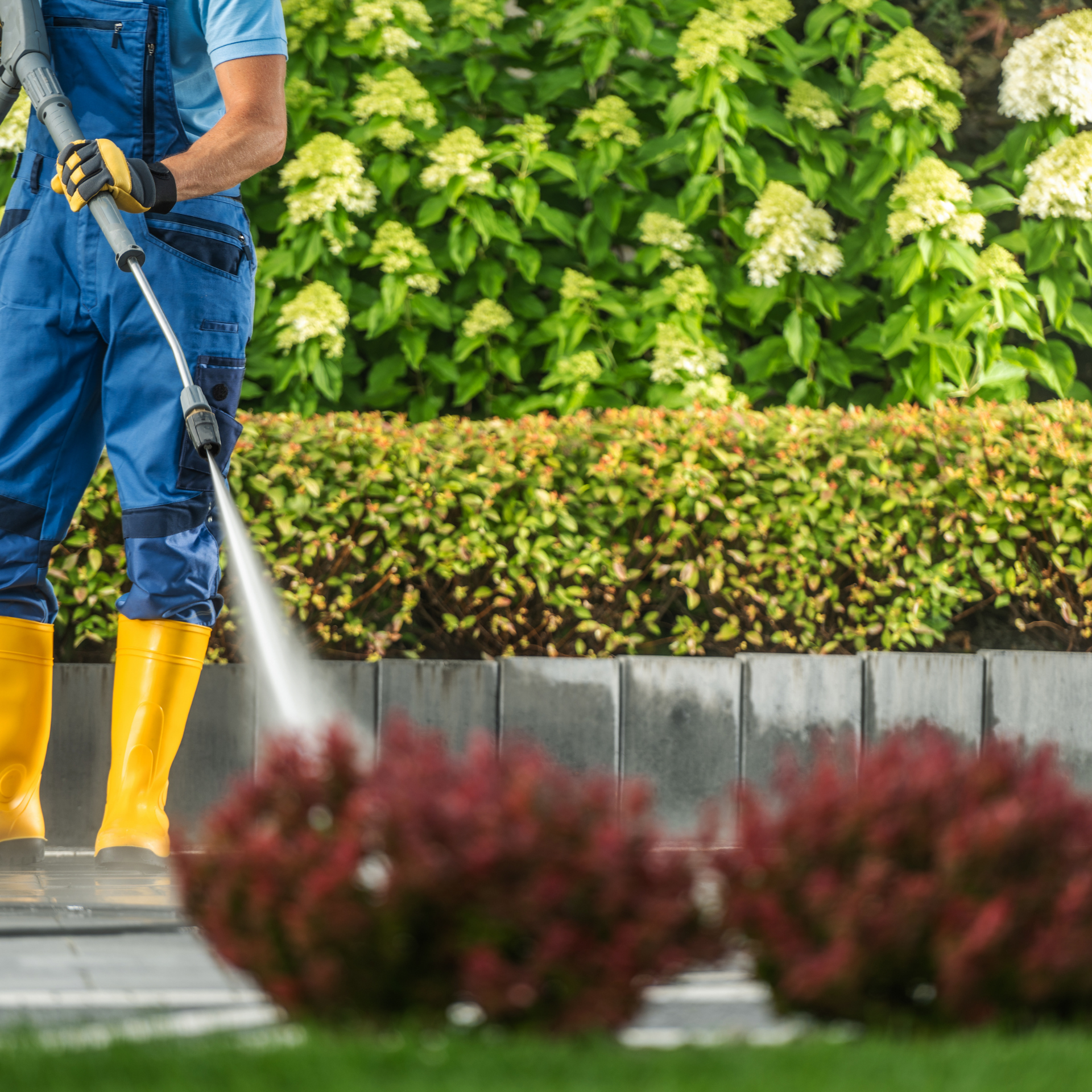 A man in blue overalls and yellow boots is using a high pressure washer to clean a sidewalk.