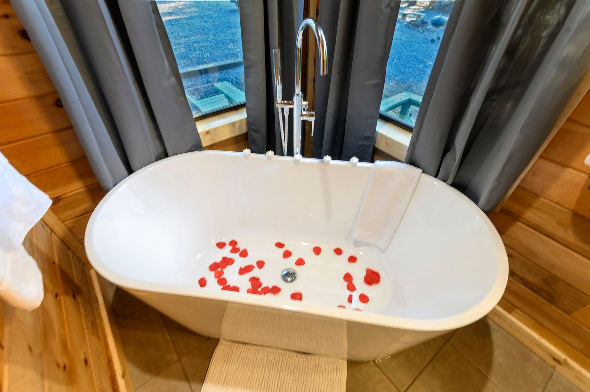 A bathtub with red petals in it is in a bathroom next to a window.