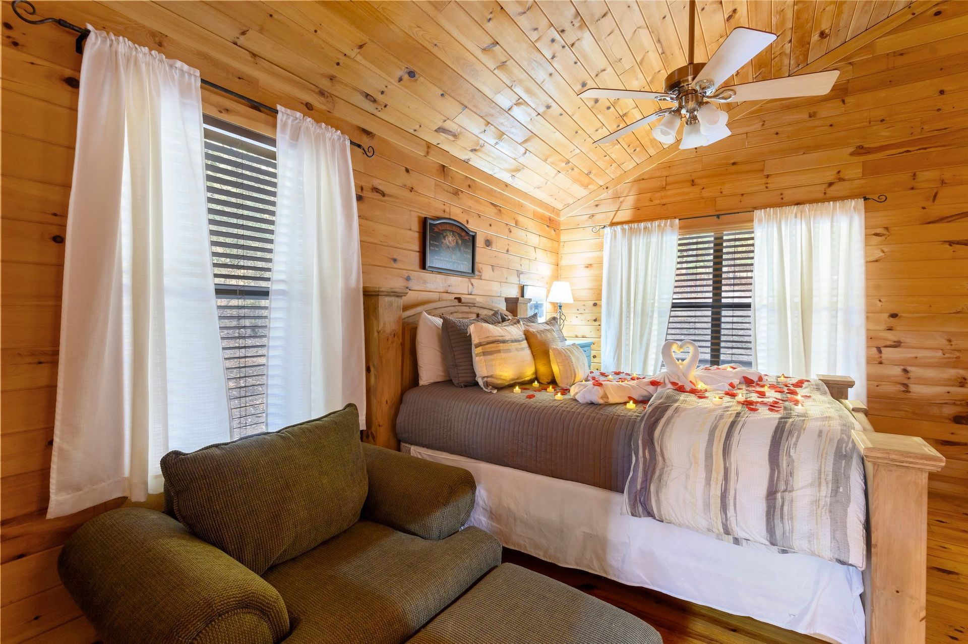 A bedroom in a log cabin with a bed , chair , ottoman and ceiling fan.
