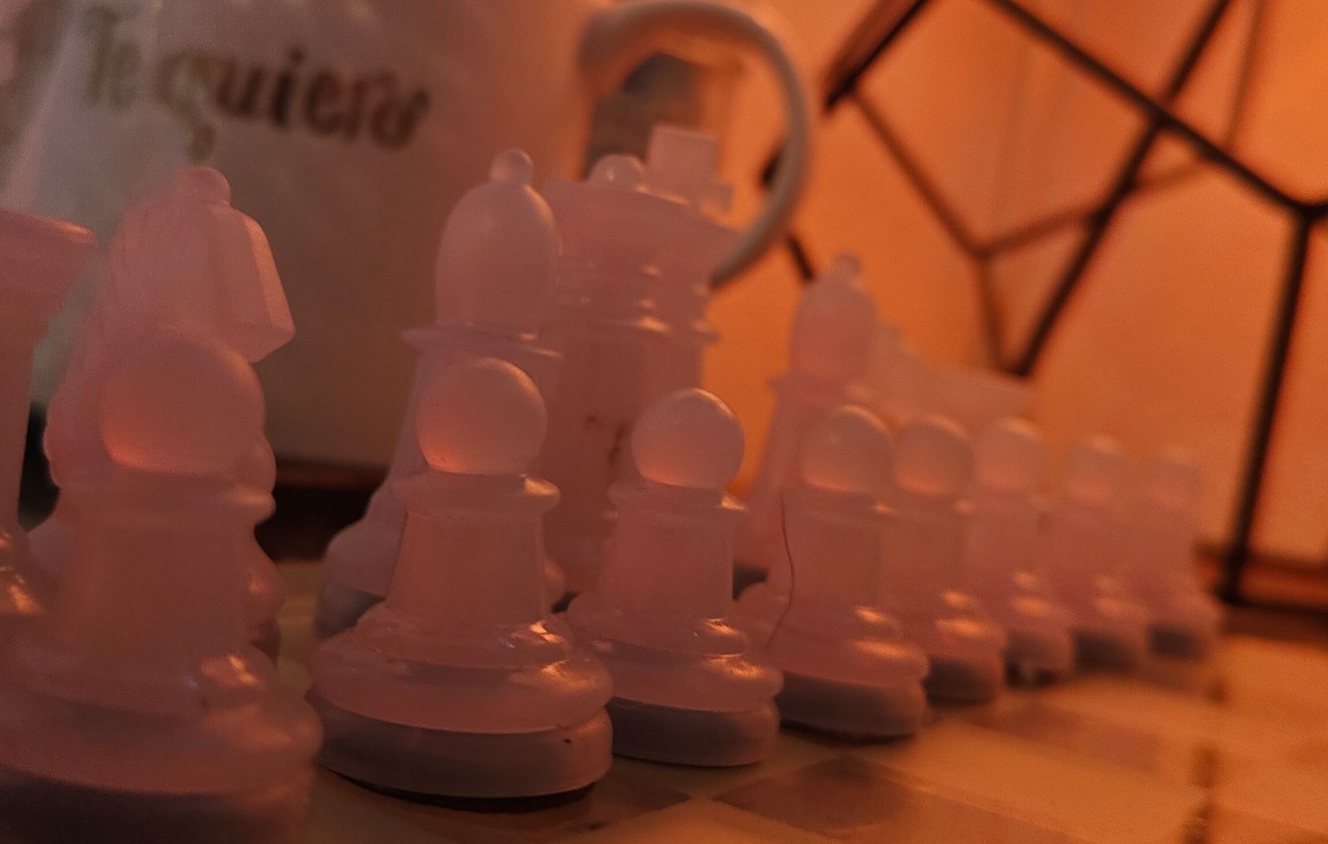 A row of translucent, pale chess pieces sits on a board in a dimly lit, warm-toned room with a mug in the background.