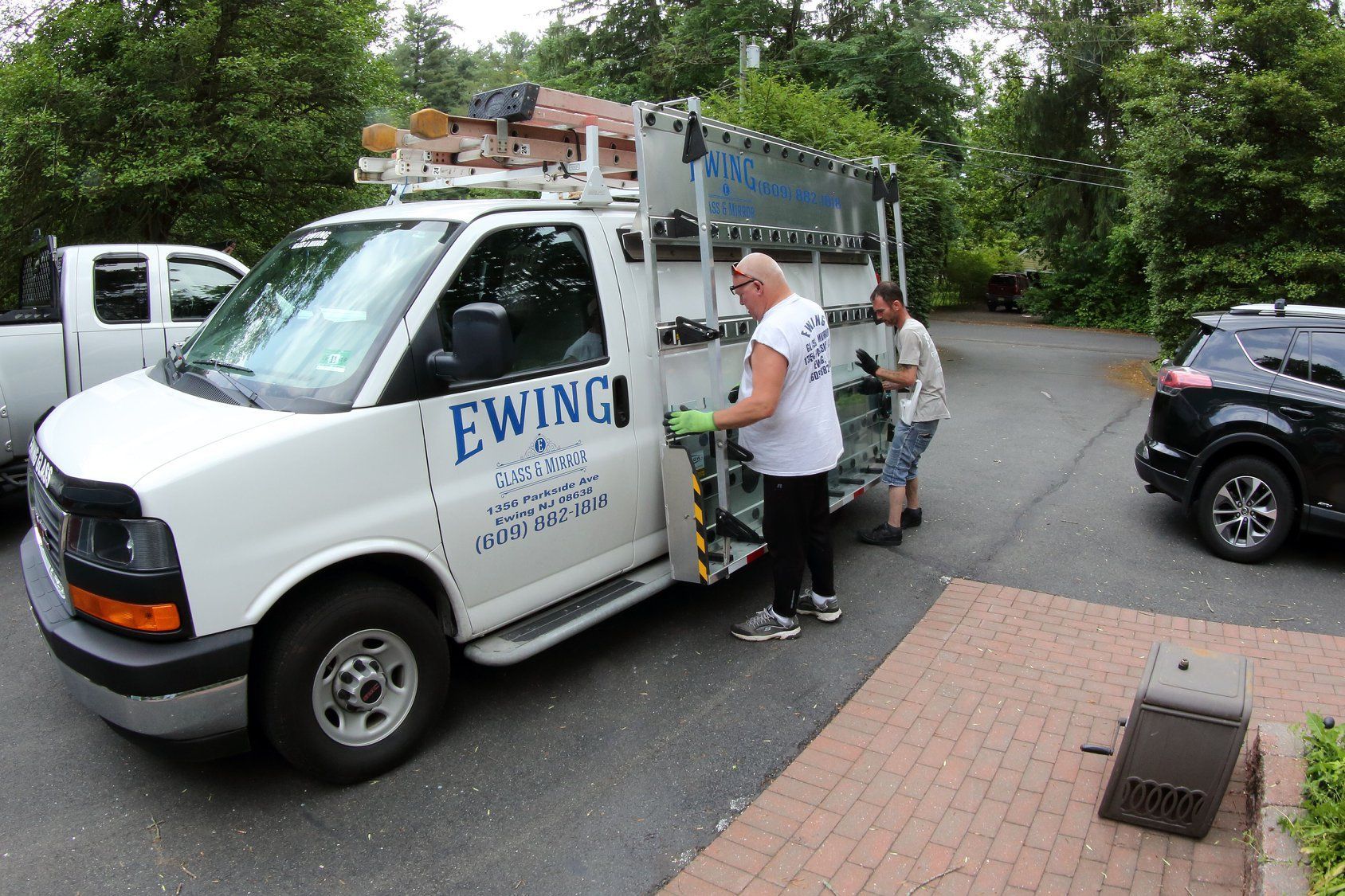 White Ewing service van with workers unloading equipment on a driveway beside a parked black SUV.
