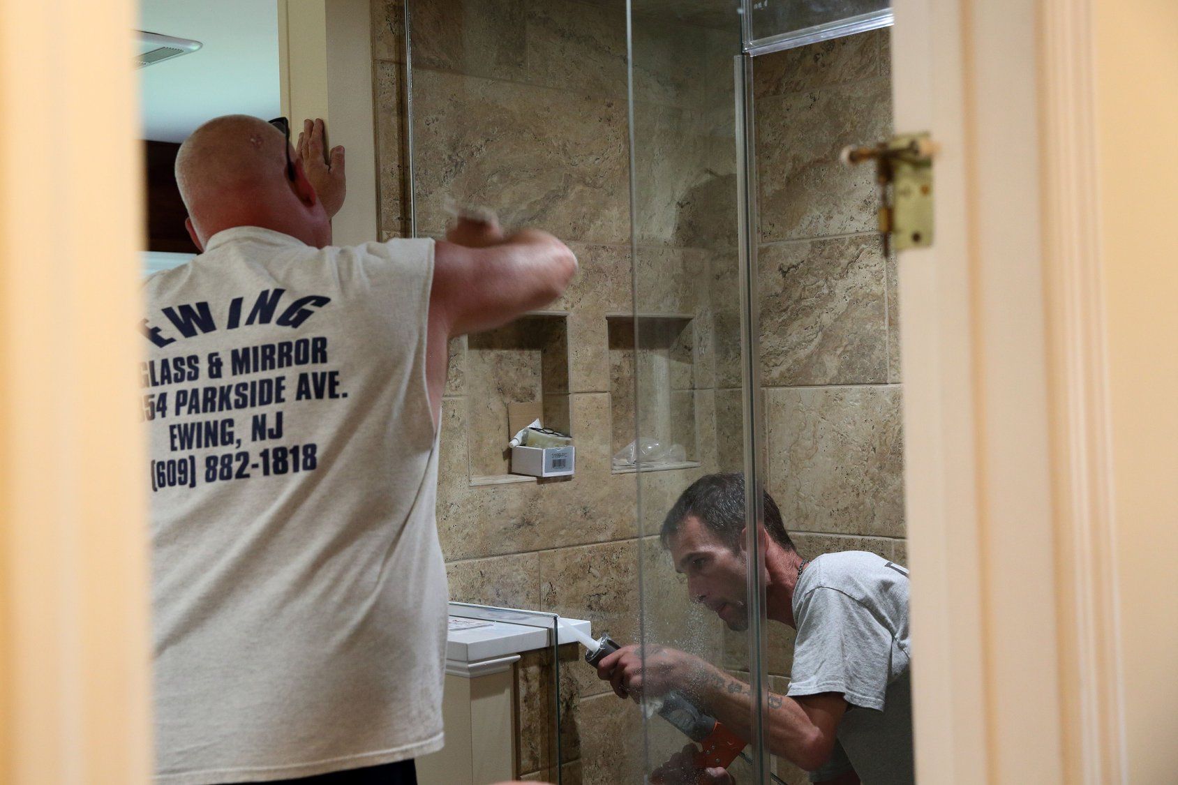 Two men working in a tiled shower stall, one installing a glass shower door