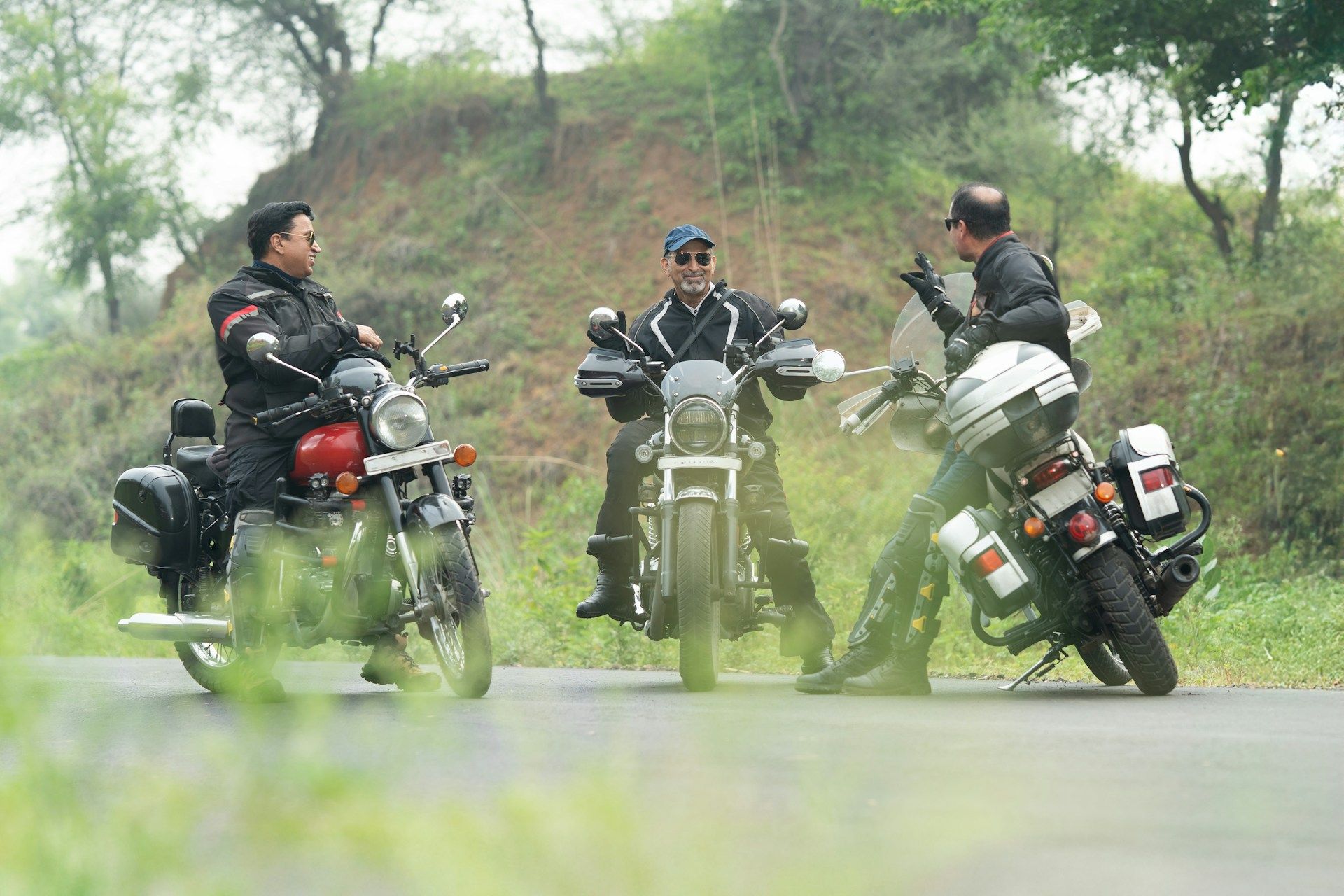 Three motorcyclists on road, stopping to chat. Two are on bikes, one gesturing. Overcast day with trees.