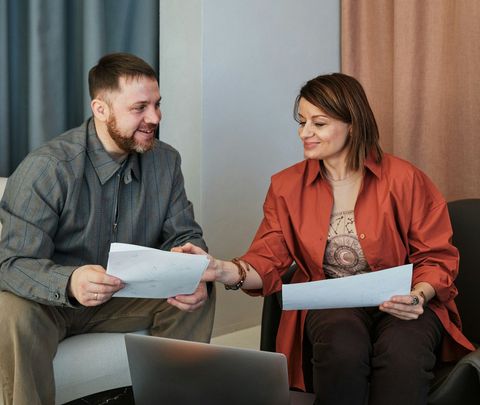 Man and woman in an office, reviewing papers. Man smiles, woman reaches to hand over a document.