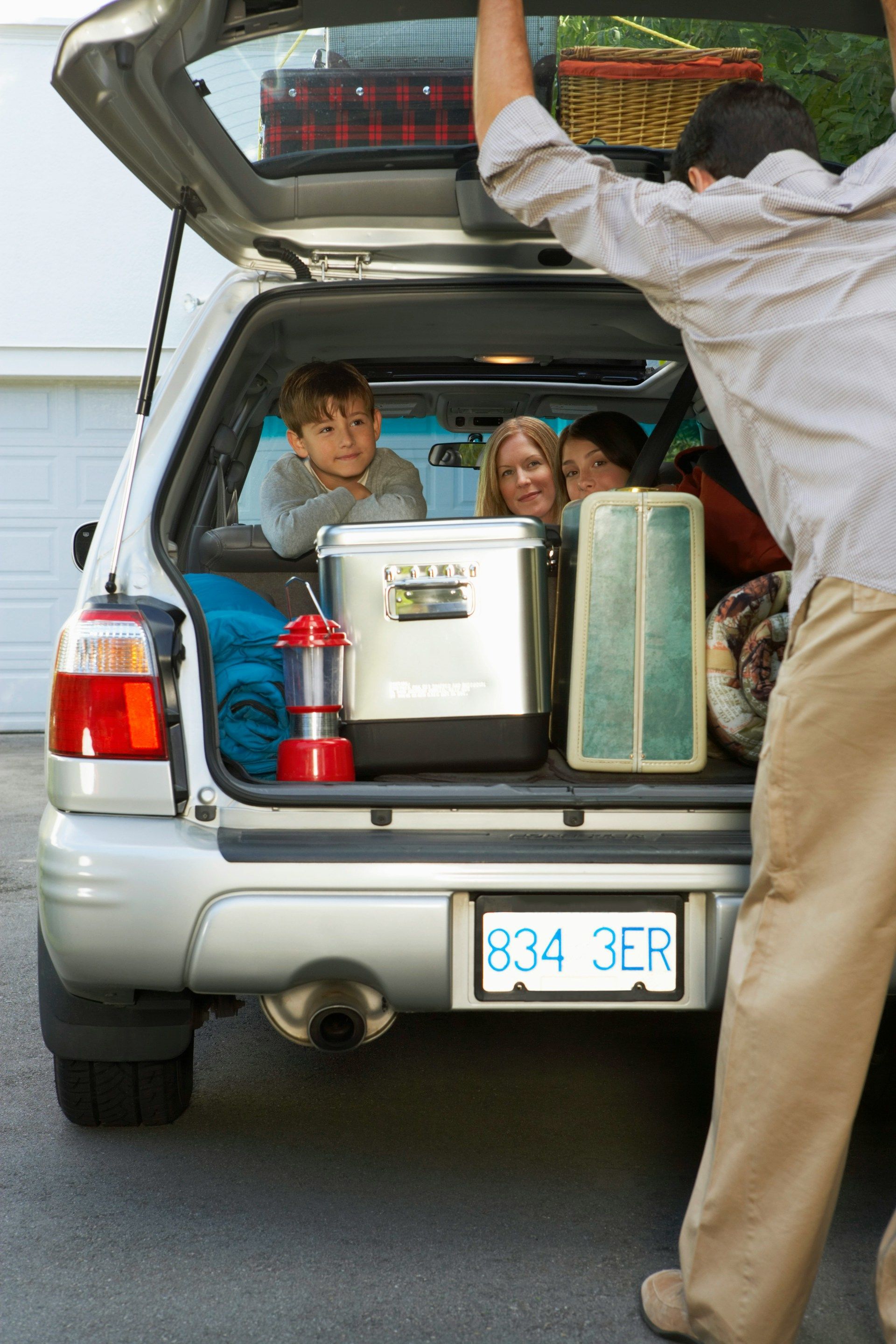 Family packing a car trunk for a trip; man loading, children looking, cooler, luggage.