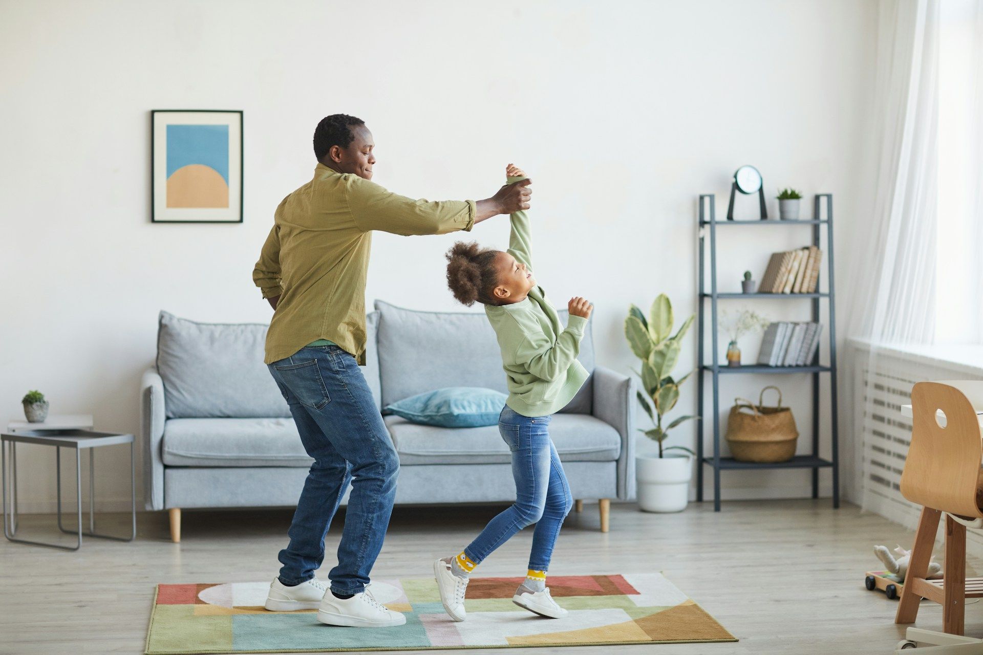 Father and daughter dance in a living room, the girl lifted by one arm.