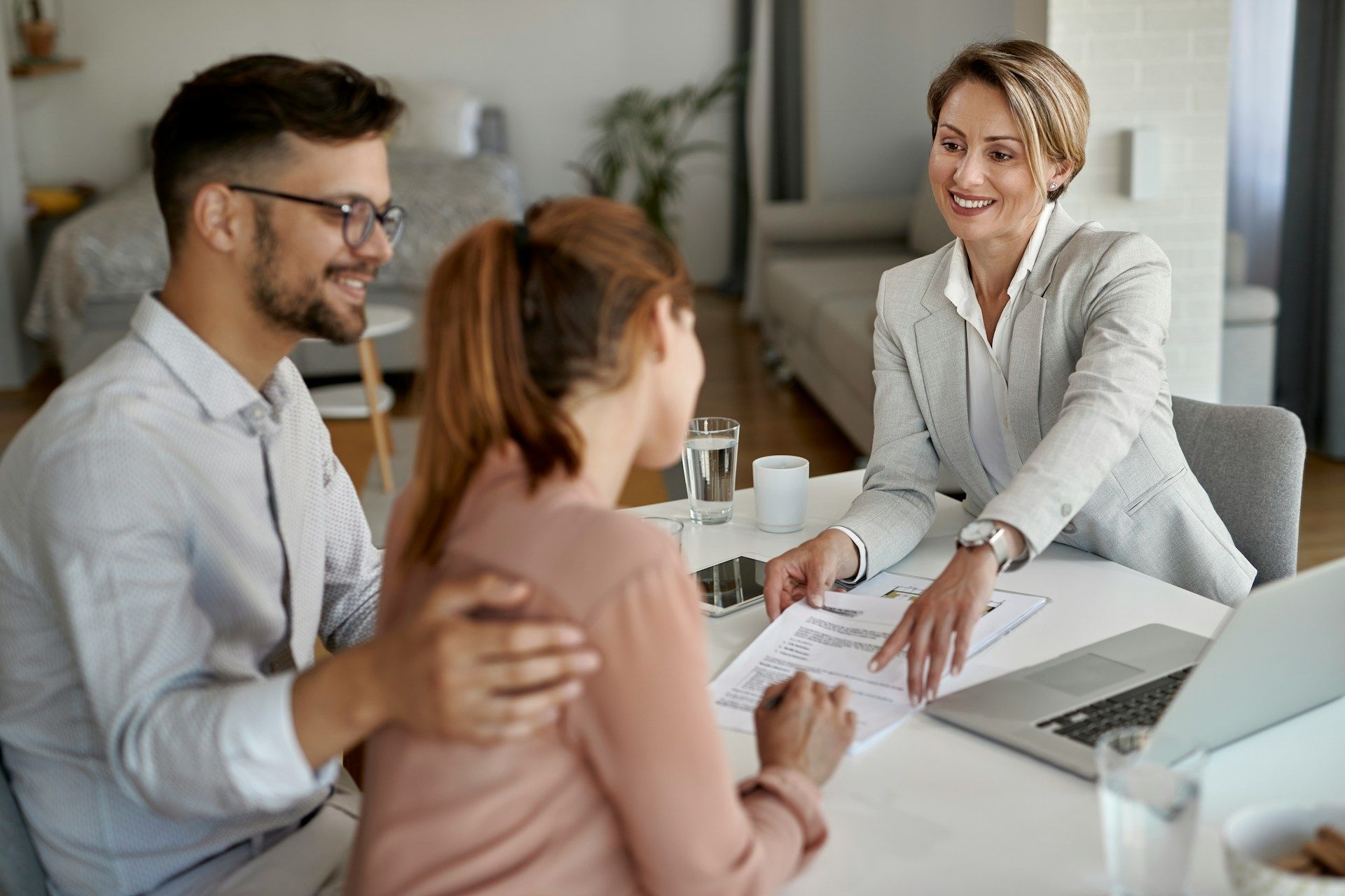 A financial advisor showing a document to a smiling couple indoors; laptop, water glass on the table.