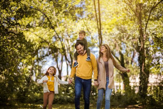 Family of four smiling and walking in a sunny park; father carries child on shoulders, holding hands with the other child and mother.