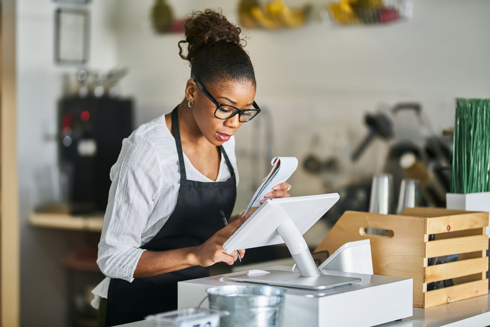 Woman with glasses and apron at a counter writing on a notepad while using a tablet.