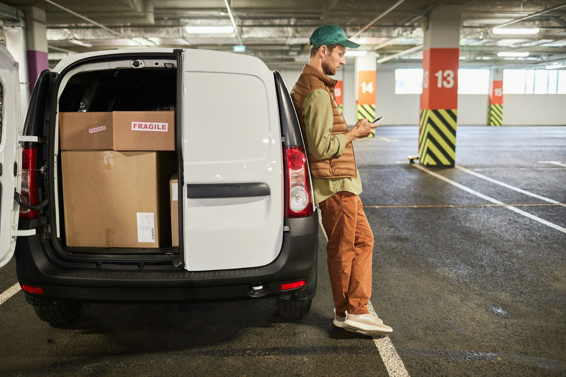 Delivery person leaning on a van in a parking garage, looking at phone, with boxes in the van.