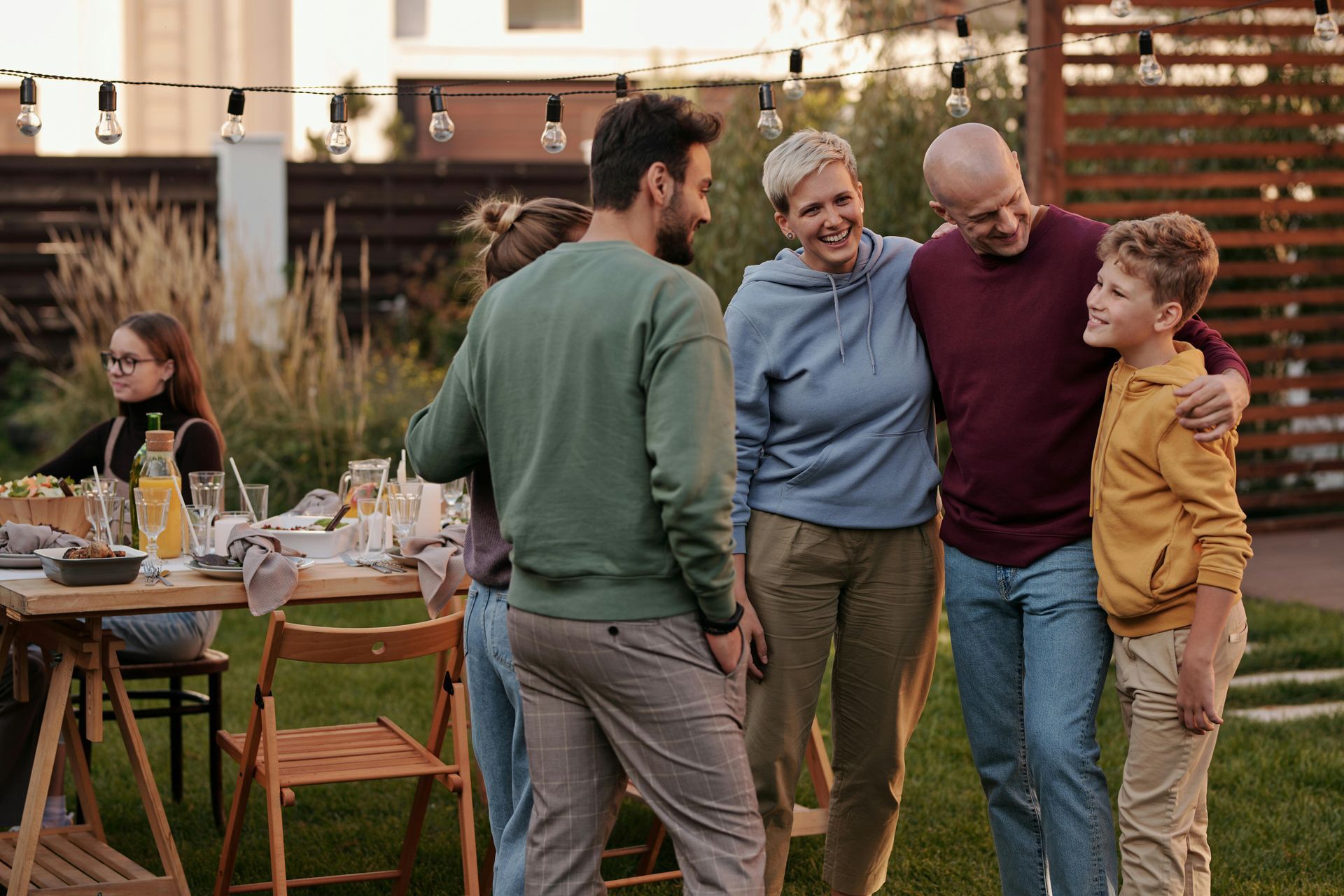 Family gathering in a backyard: people smiling, some with arms around each other, dining table set with food and drinks.