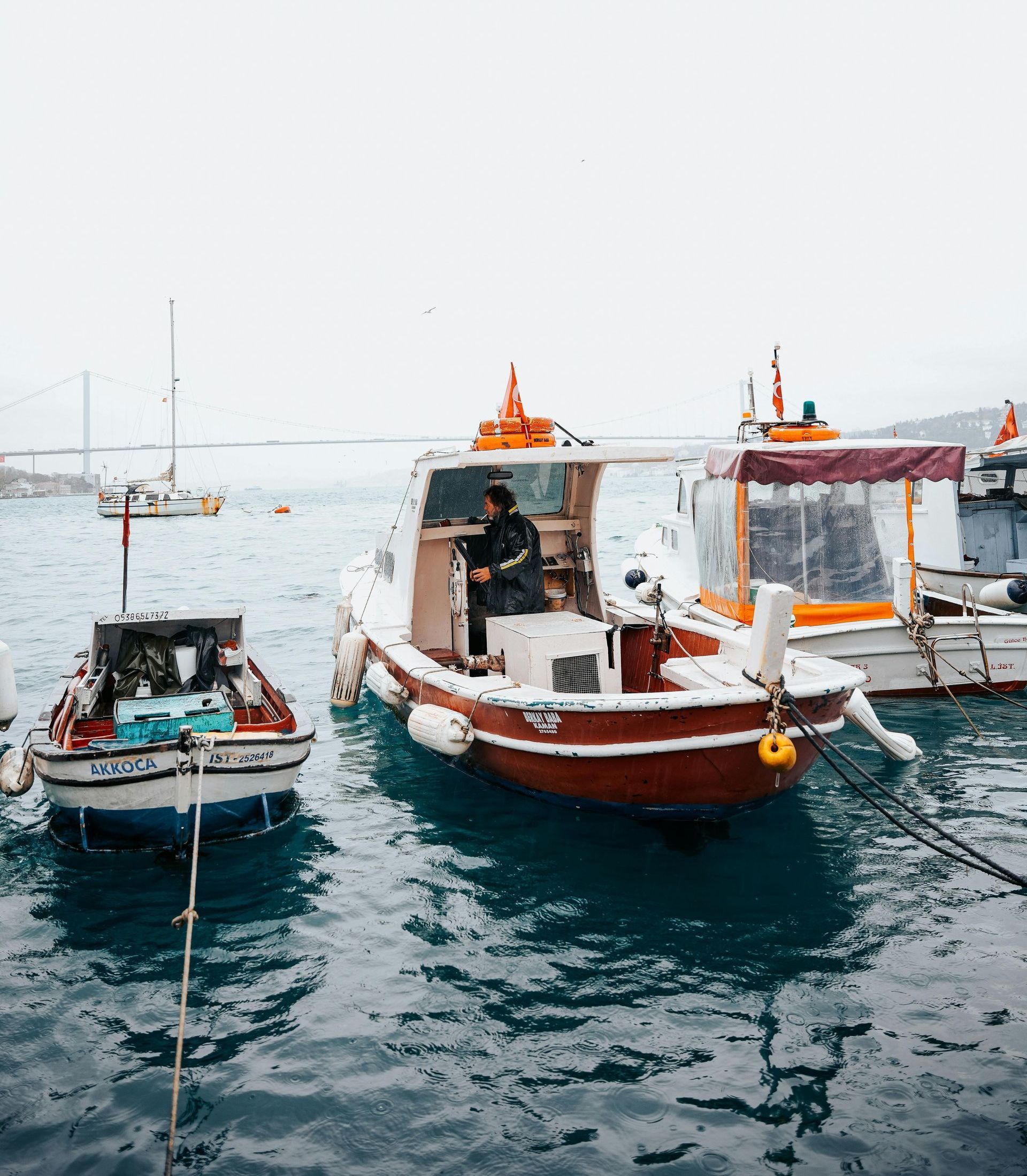 Boats docked in blue water on an overcast day; a person steers a red and white boat.