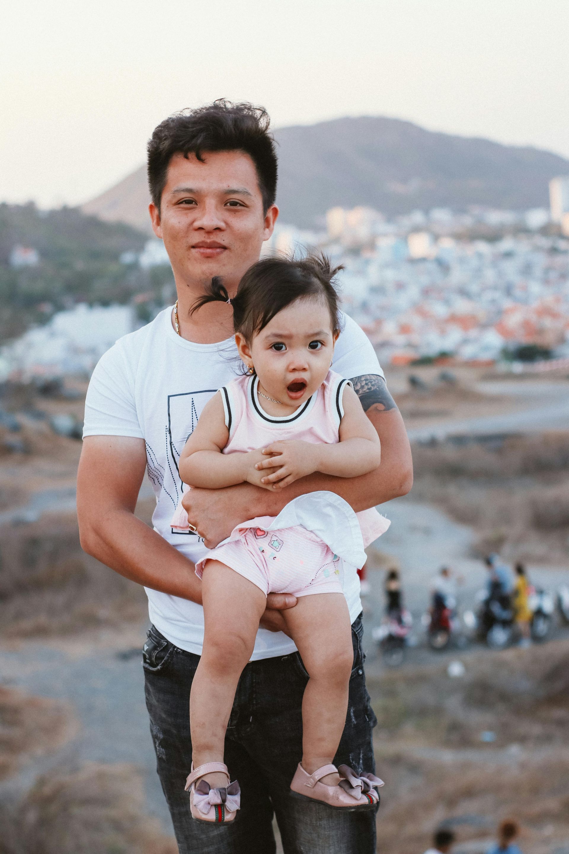 Man holding a baby with an open mouth, standing on a hill with a city and mountains in the background.