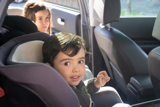 Child in car seat looking at camera, another child in background.