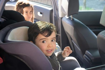 Child in car seat looking at camera, another child in background.