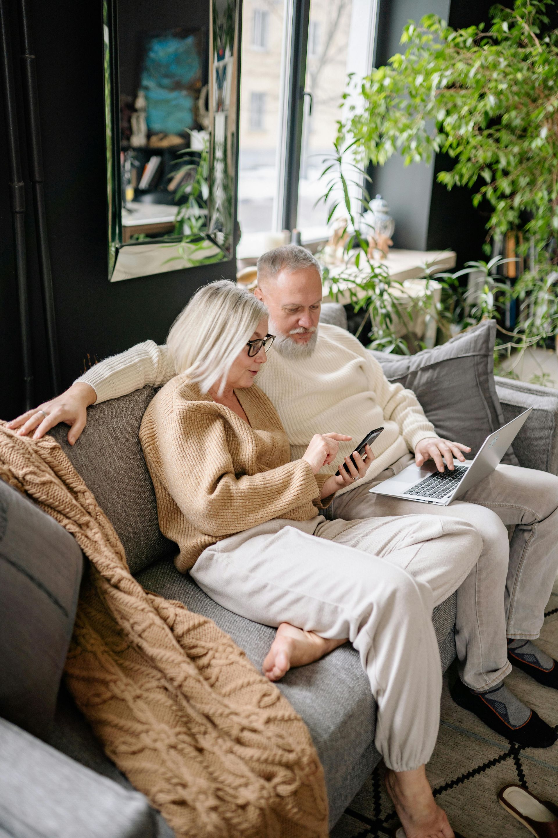Elderly couple on couch: woman with phone, man with laptop, near window.