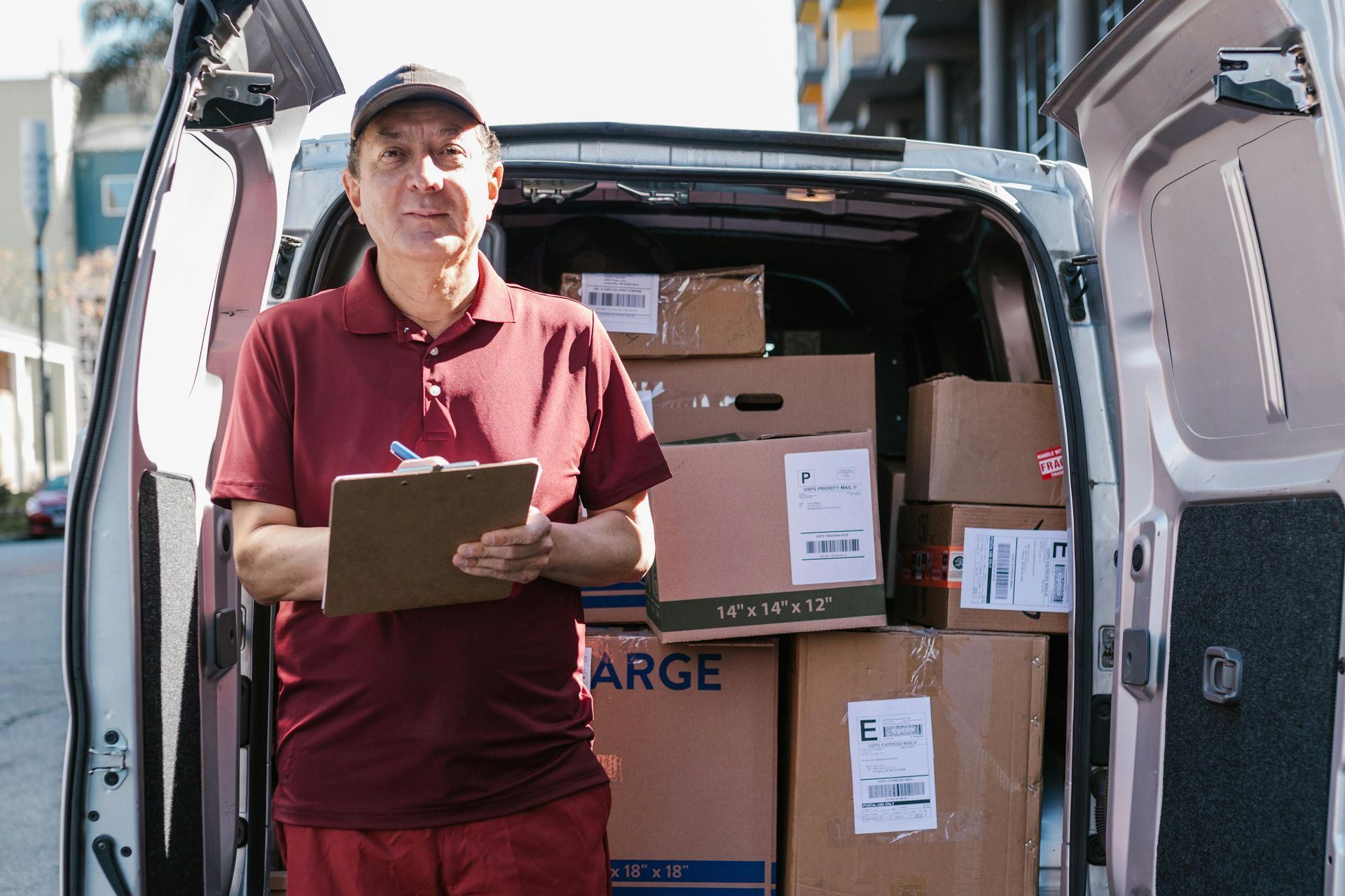 Delivery person in a maroon shirt and cap, smiling, holding a clipboard, standing in back of delivery van filled with boxes.
