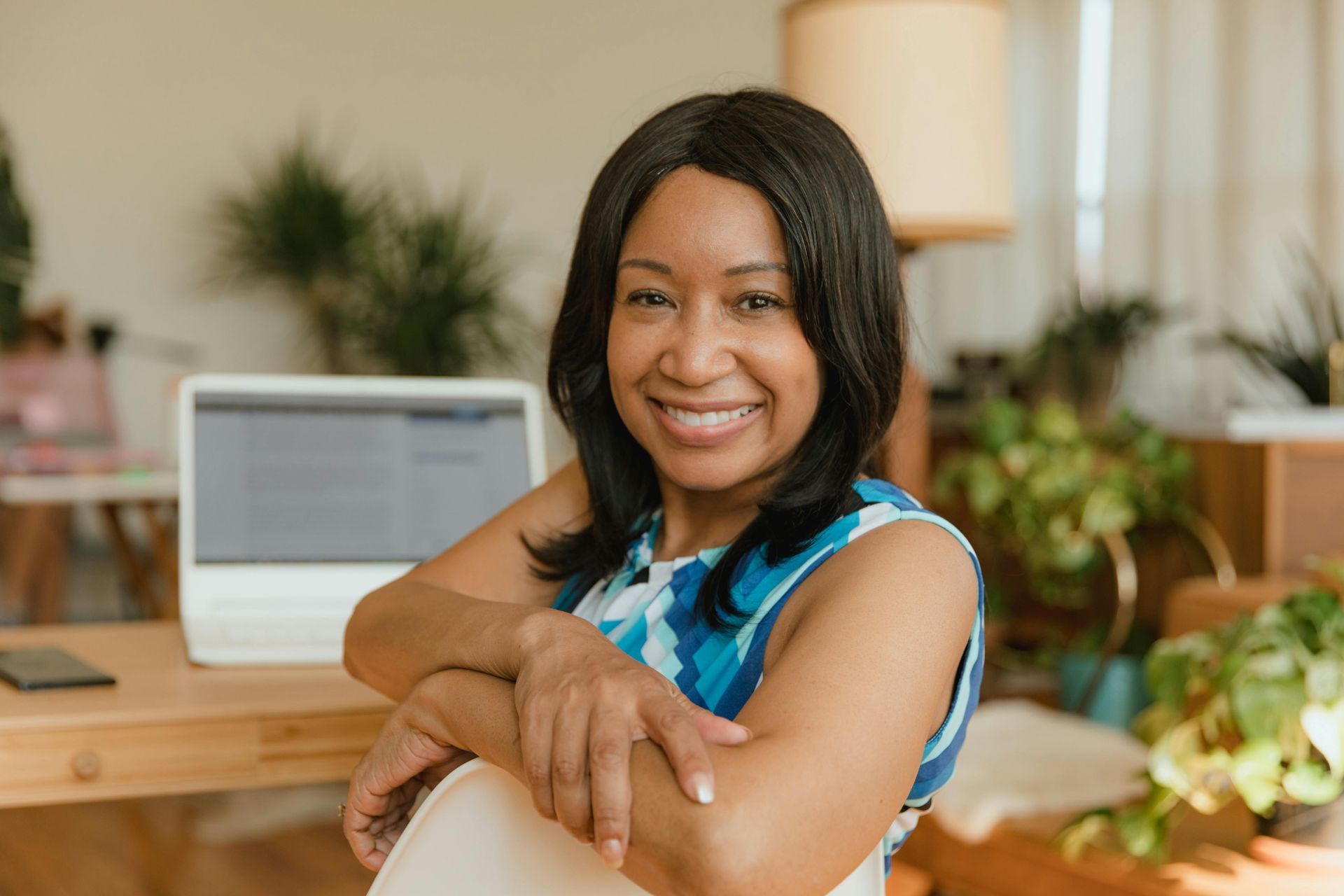 Smiling woman at desk with laptop, surrounded by plants. She's wearing a blue patterned top.