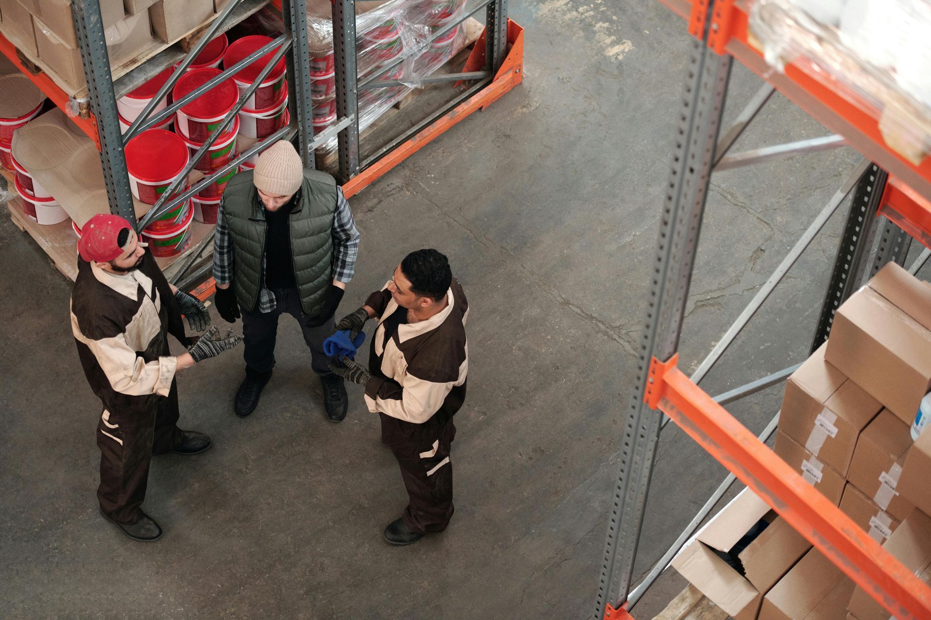 Three workers in a warehouse, one with a jacket and two in jumpsuits, talking near shelves.