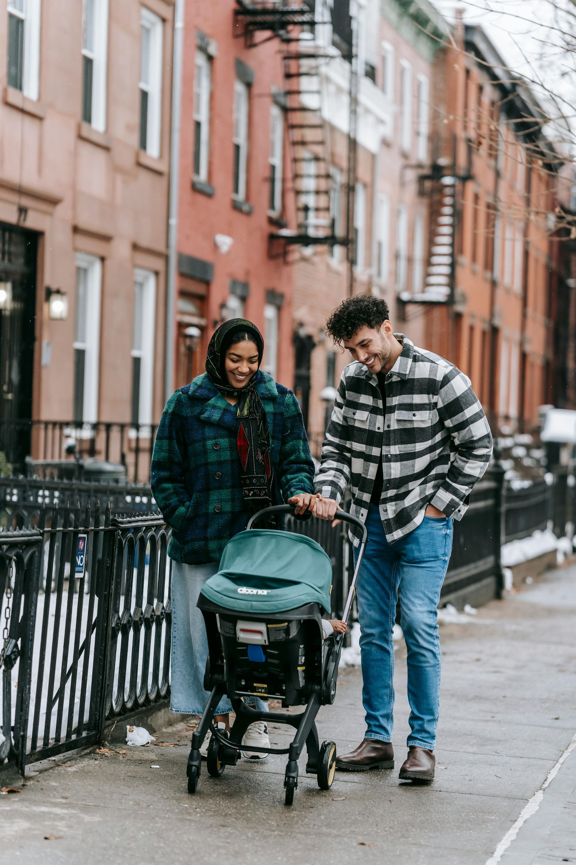 Couple walking with a stroller on a city sidewalk. Man looking down, woman smiling. Buildings in background.
