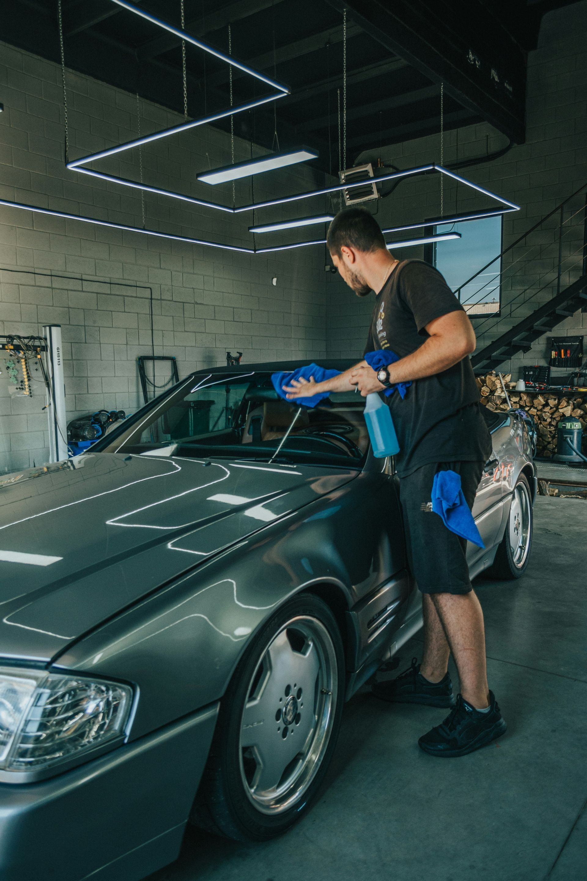 Man cleaning a silver car's windshield with blue cloth and spray bottle in a garage under neon lights.