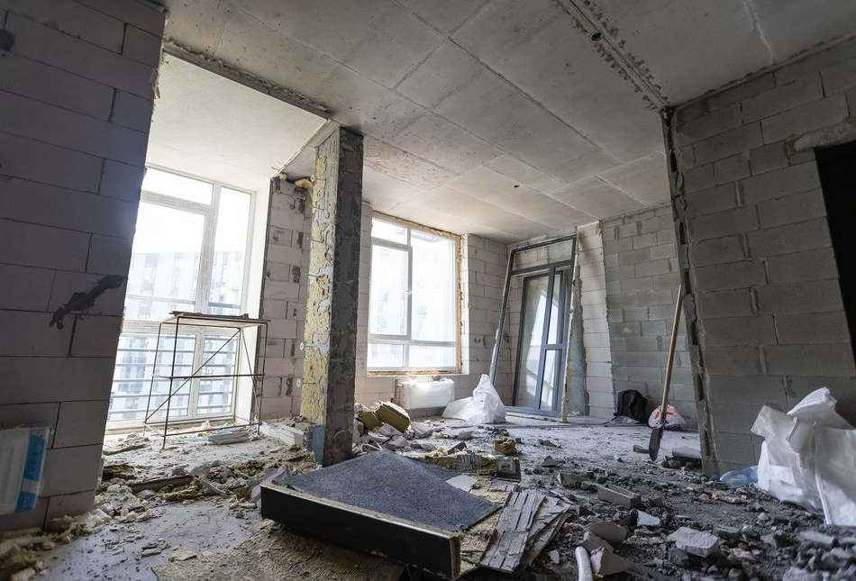 Empty, unfinished room with exposed cinderblock walls, debris on the floor, and large windows letting in daylight