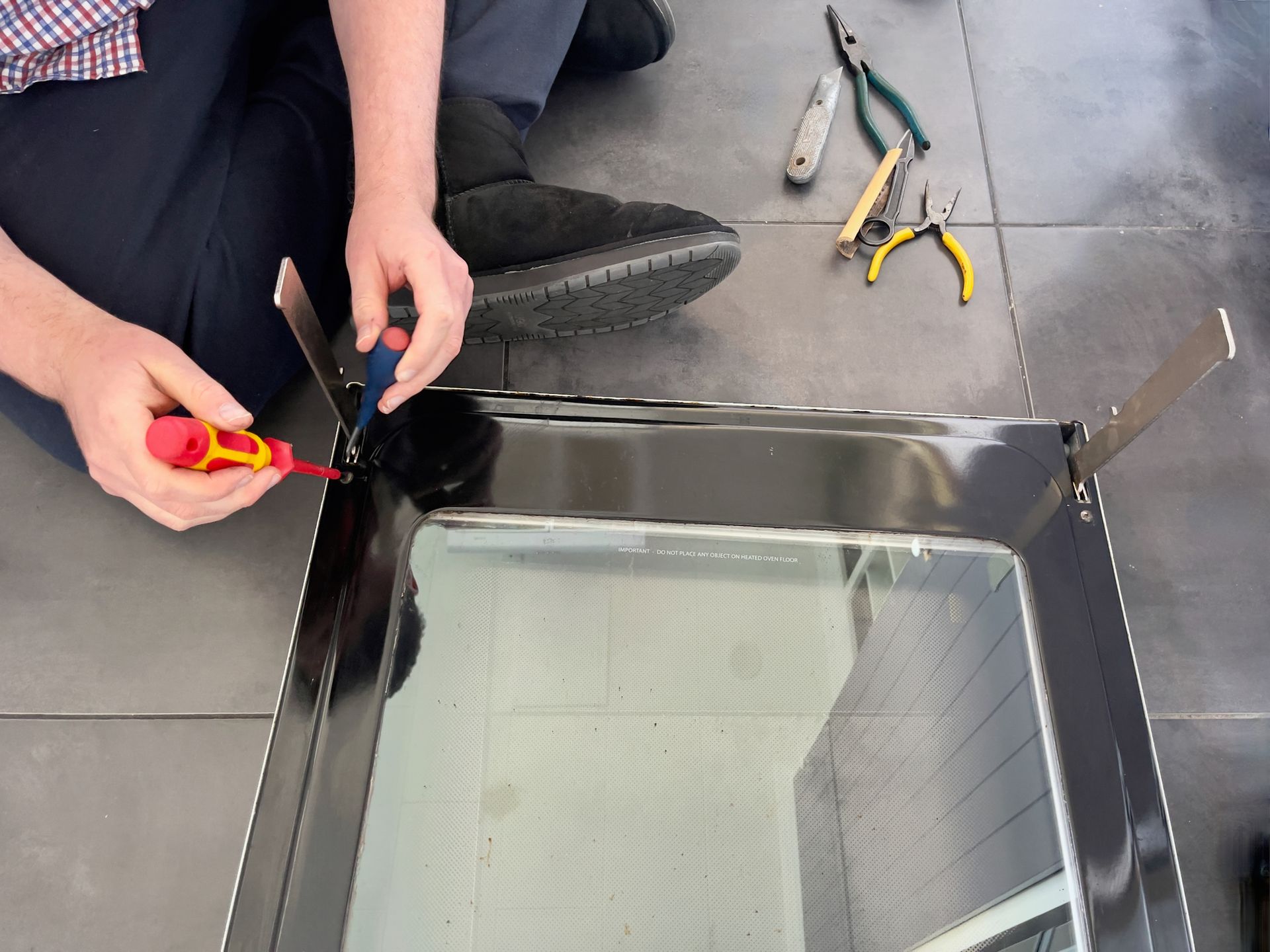 A man is kneeling on the floor fixing an oven door with a screwdriver.