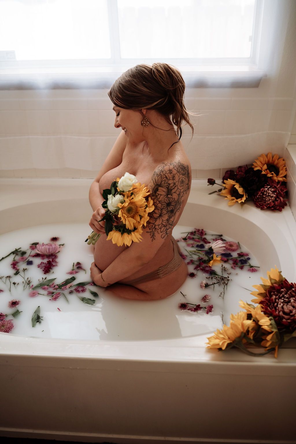 A pregnant woman is kneeling in a bathtub filled with milk and flowers.