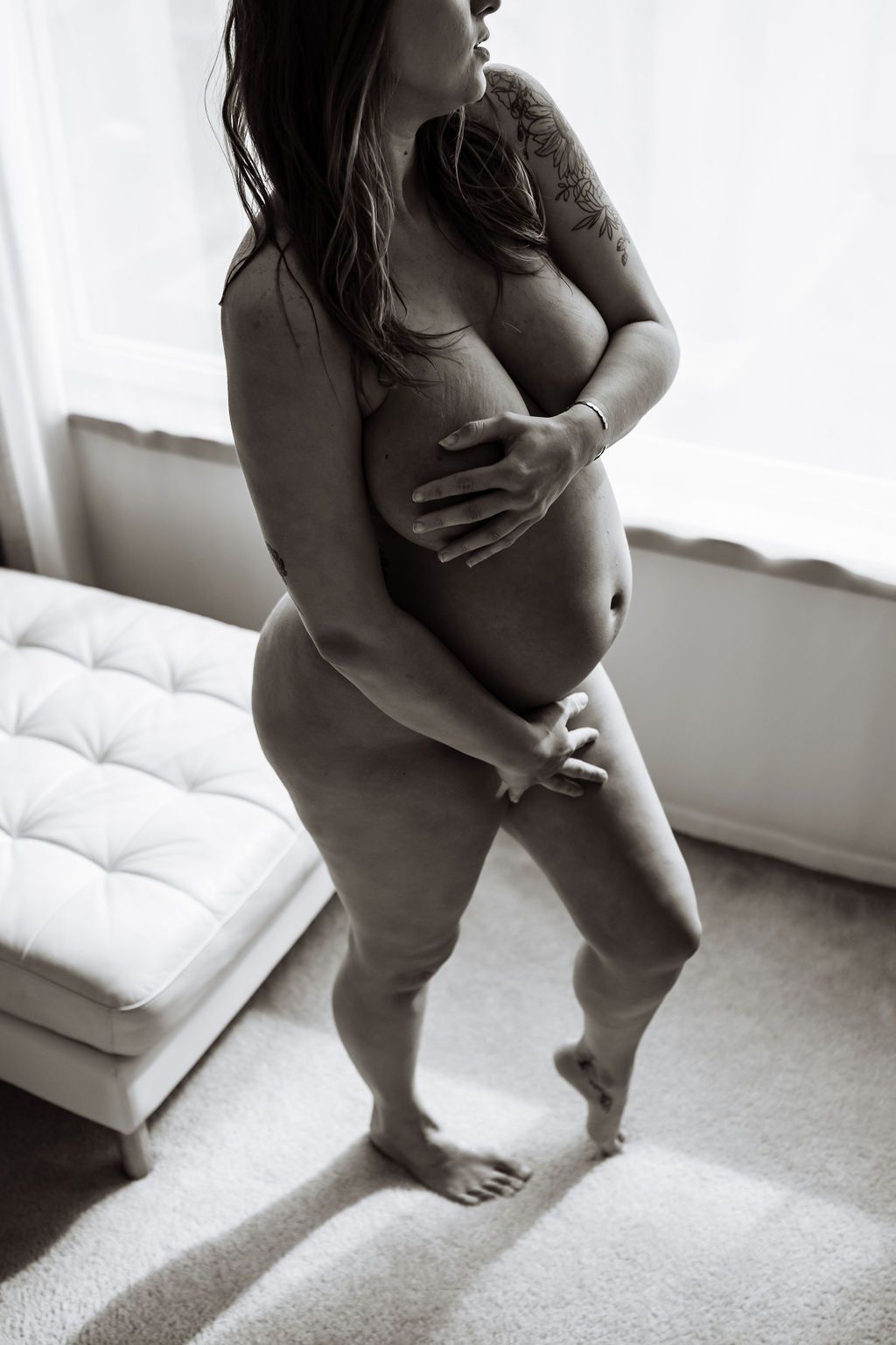 A nude pregnant woman standing in front of a window with her hands around her belly and covering her breasts 