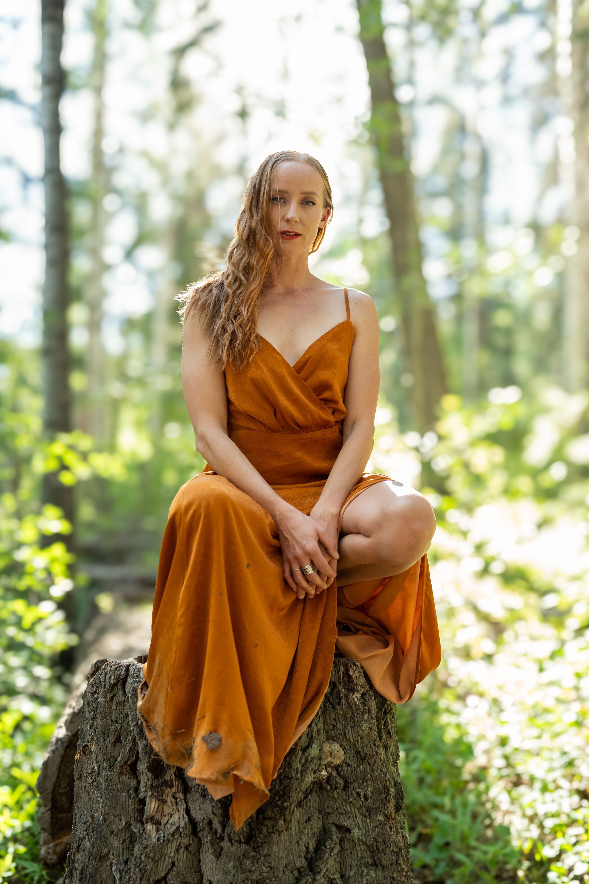 A woman in a long orange dress is sitting on a tree stump in the woods.