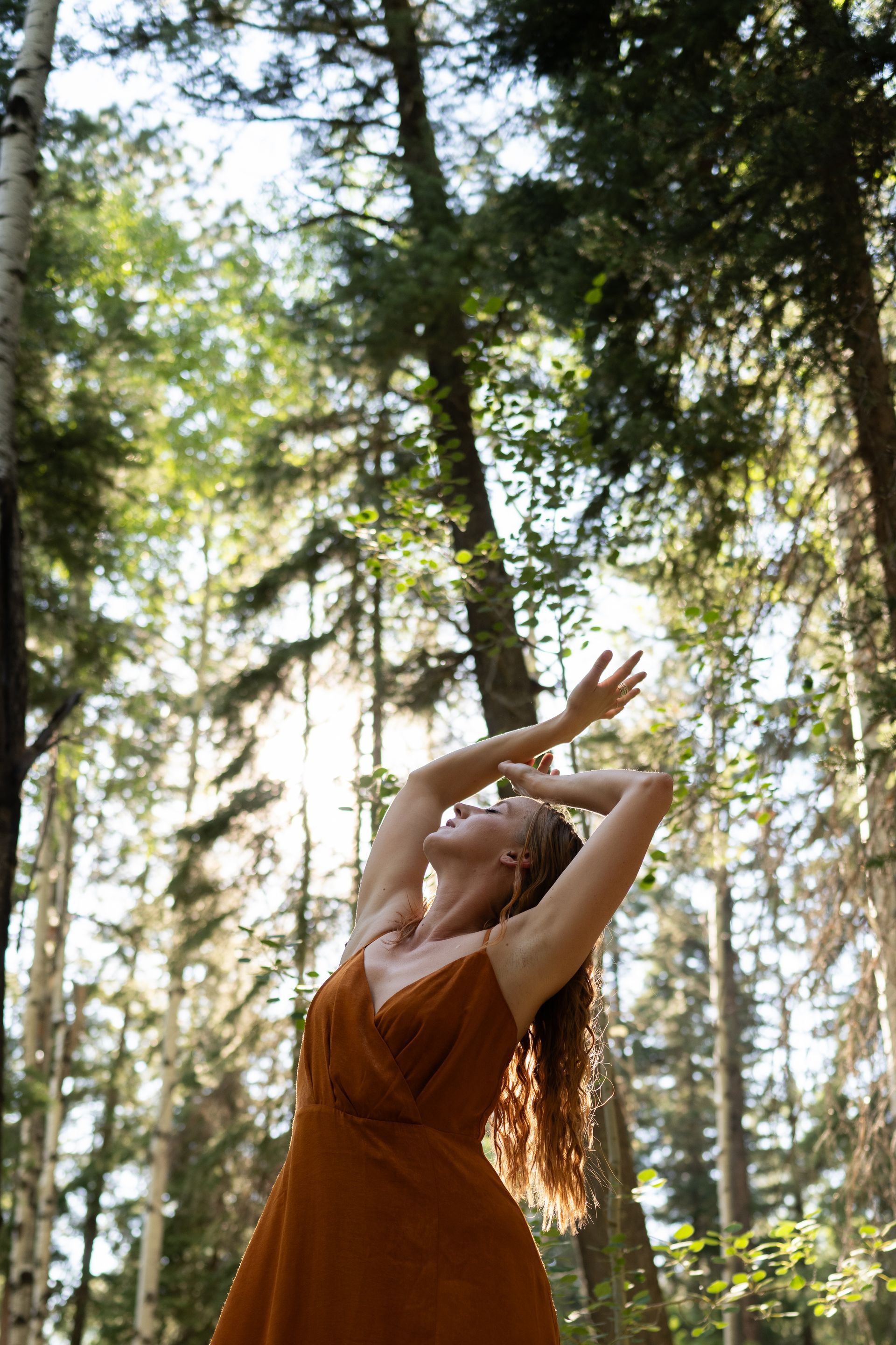 A woman in a dress is standing in the middle of a forest with her arms outstretched.