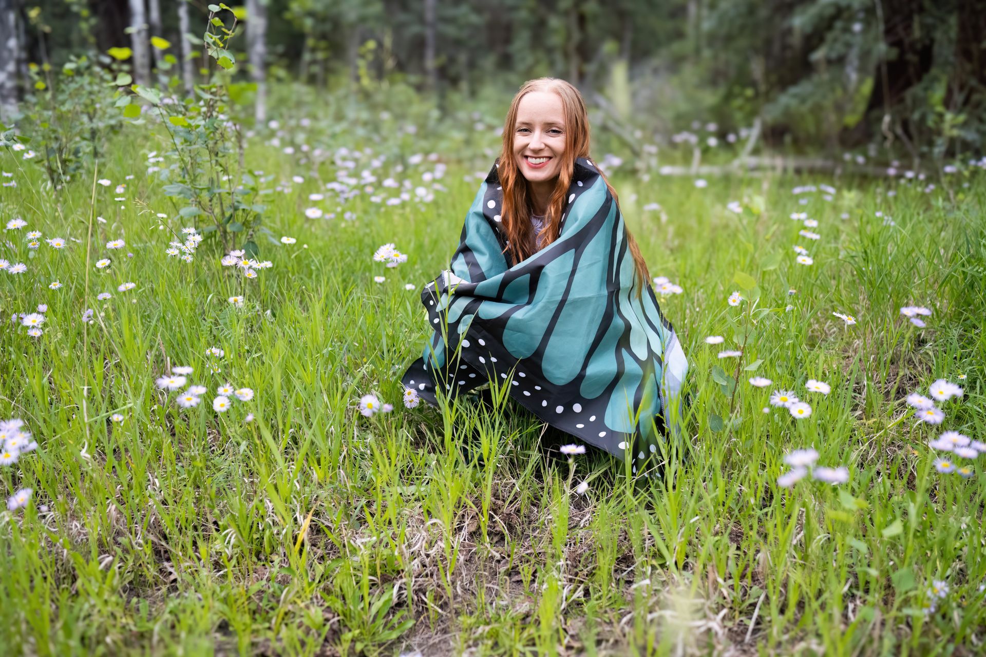 A woman is sitting in a field of flowers wrapped in a butterfly blanket.