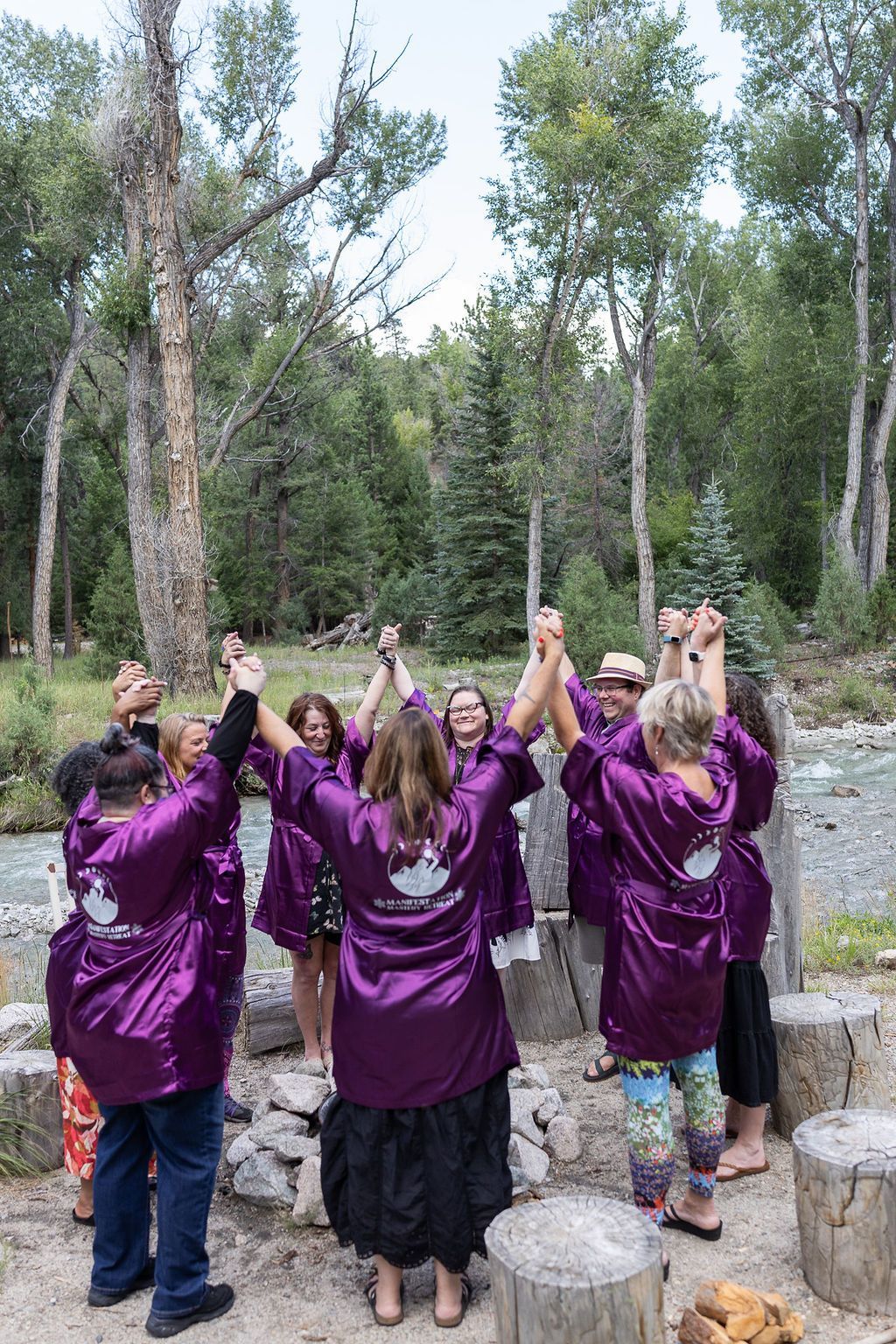 Group in purple robes holding hands in a circle, near a stream and trees, outside.