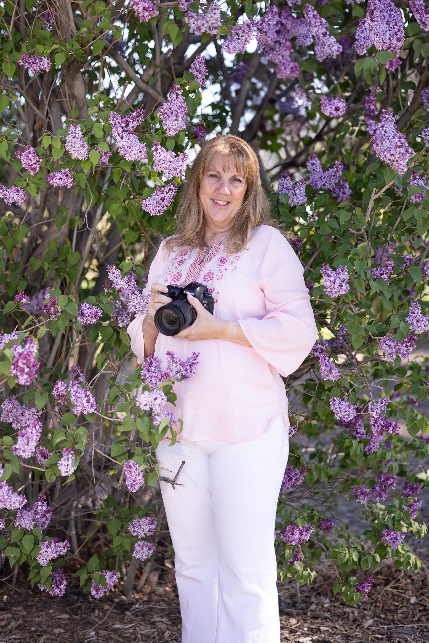Woman holding camera, smiles, stands amidst blooming lilac bush.