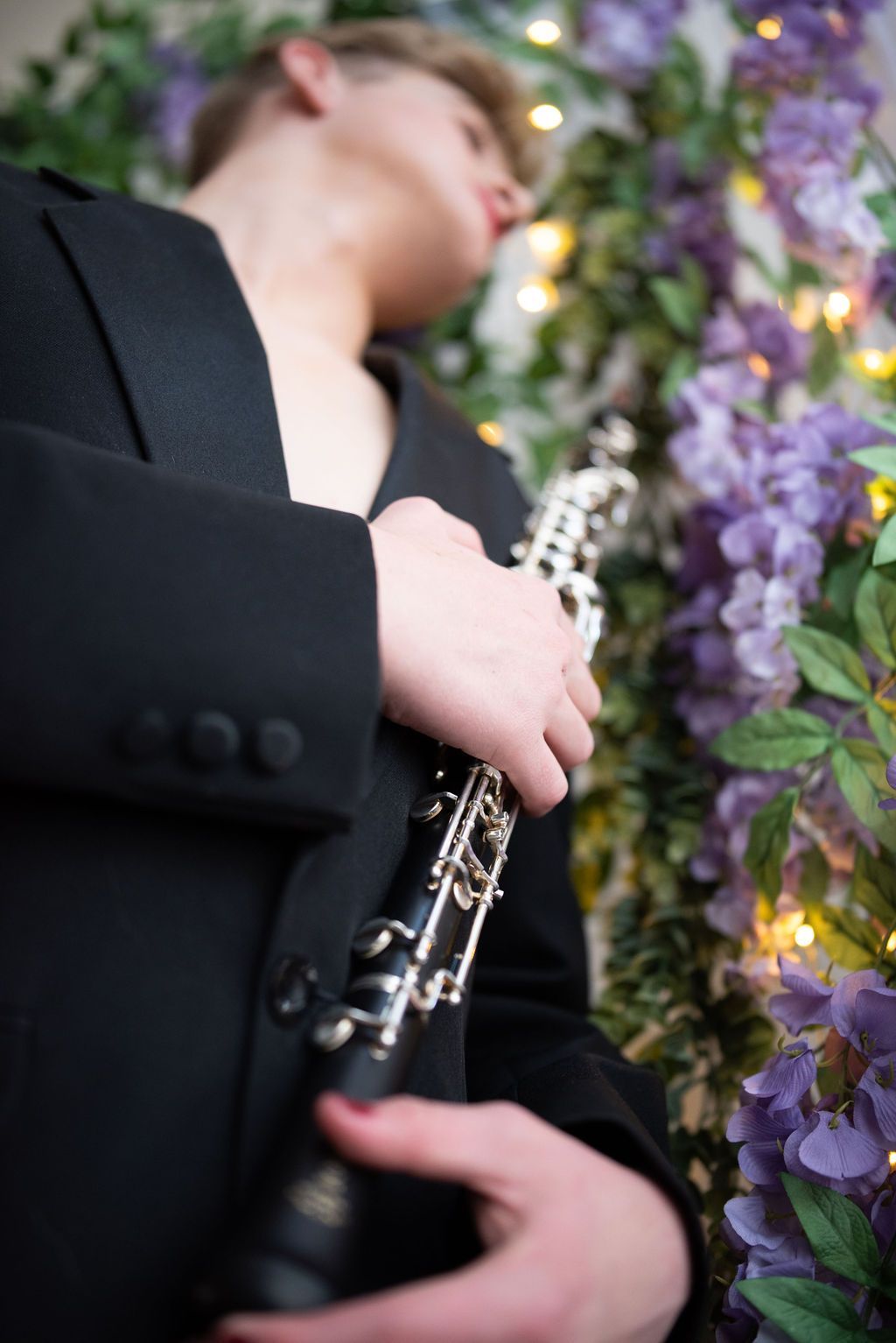 A man in a suit is playing a clarinet in front of purple flowers.