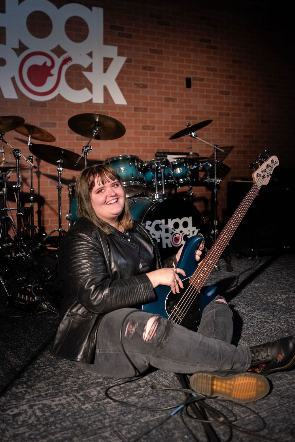A woman is sitting on the floor holding a guitar in front of a drum set.