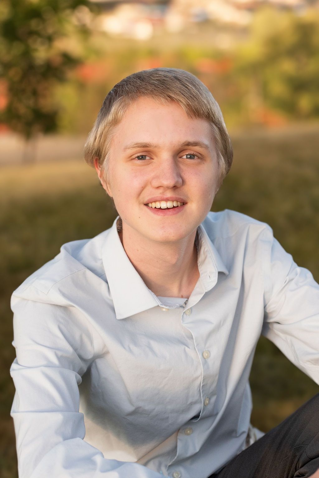A young man in a white shirt is sitting in the grass and smiling for the camera.