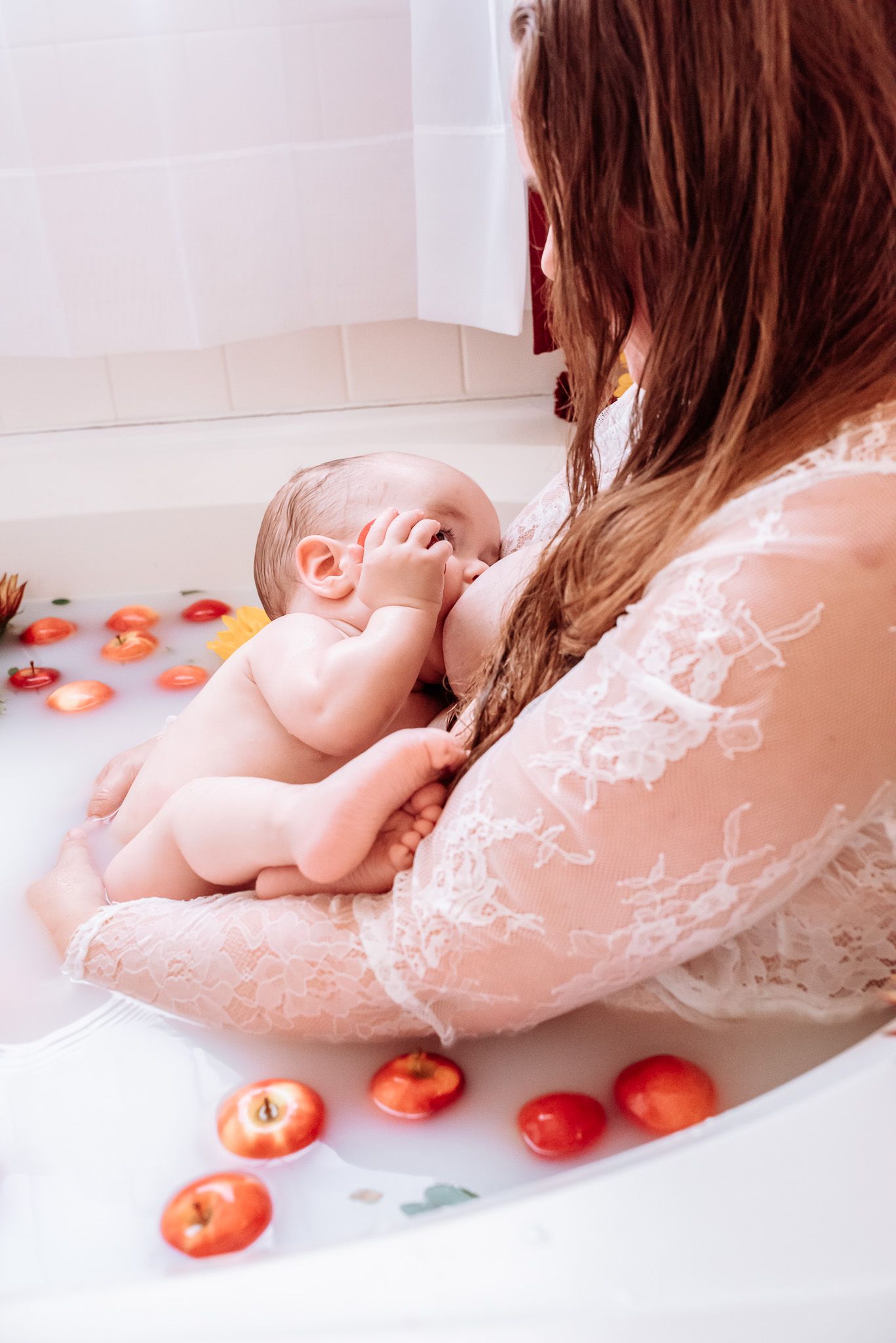 A woman is breastfeeding a baby in a bathtub filled with milk and apples.