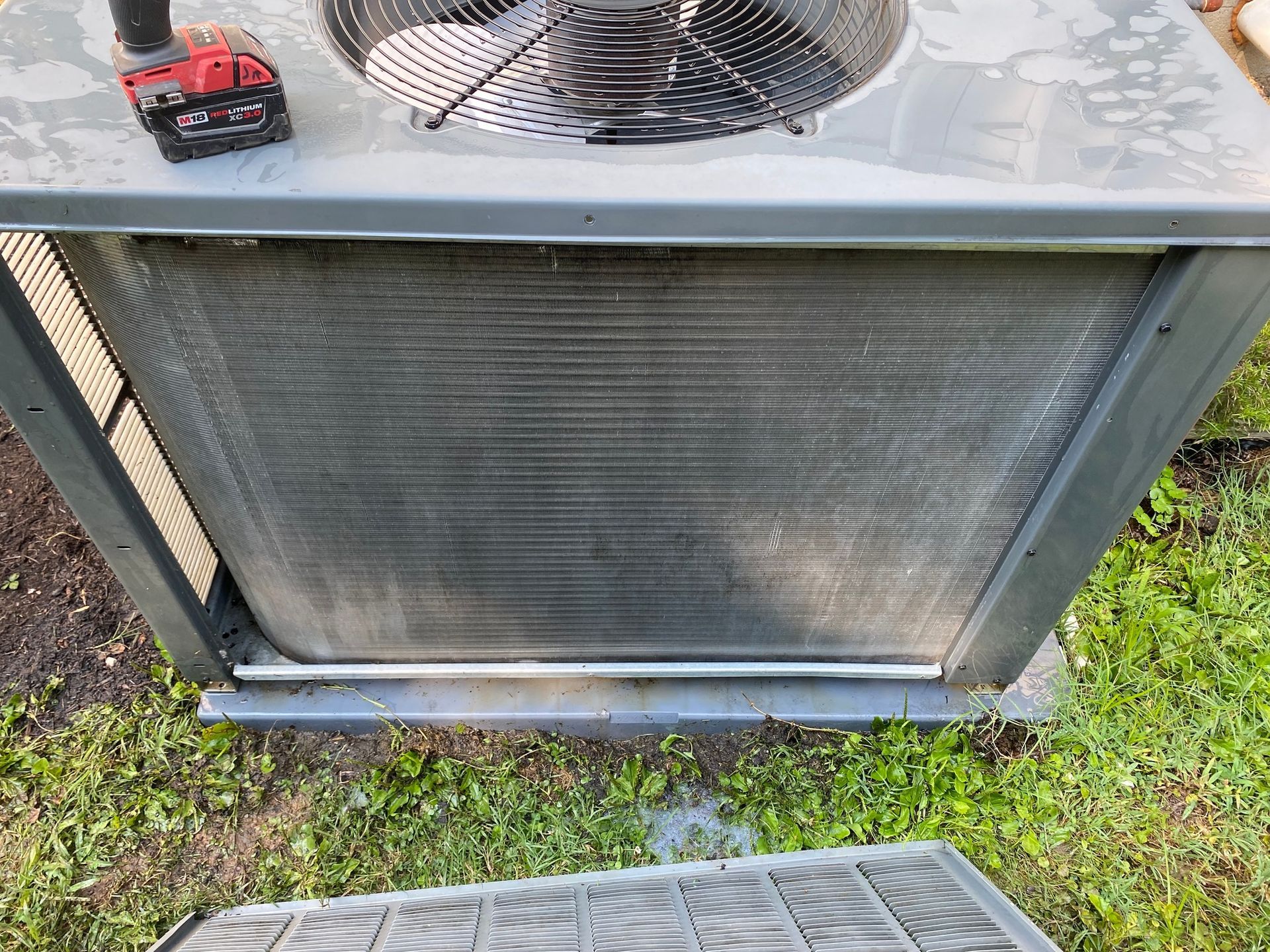 A dirty air conditioner is sitting in the grass next to a drill.
