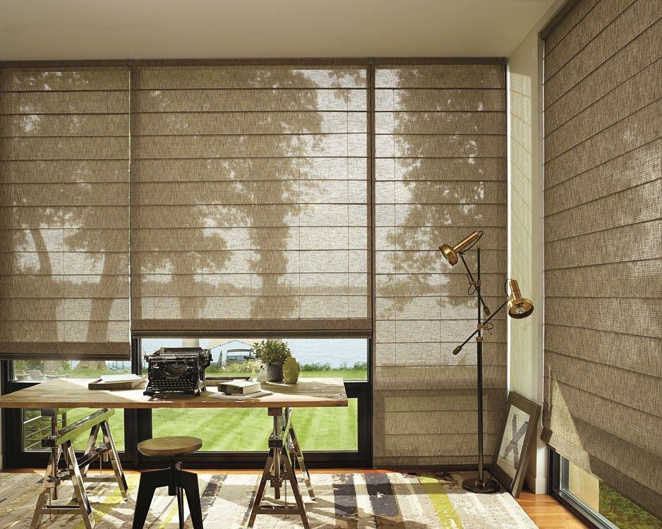 A room with a desk , chair , lamp and a large window with roman shades.