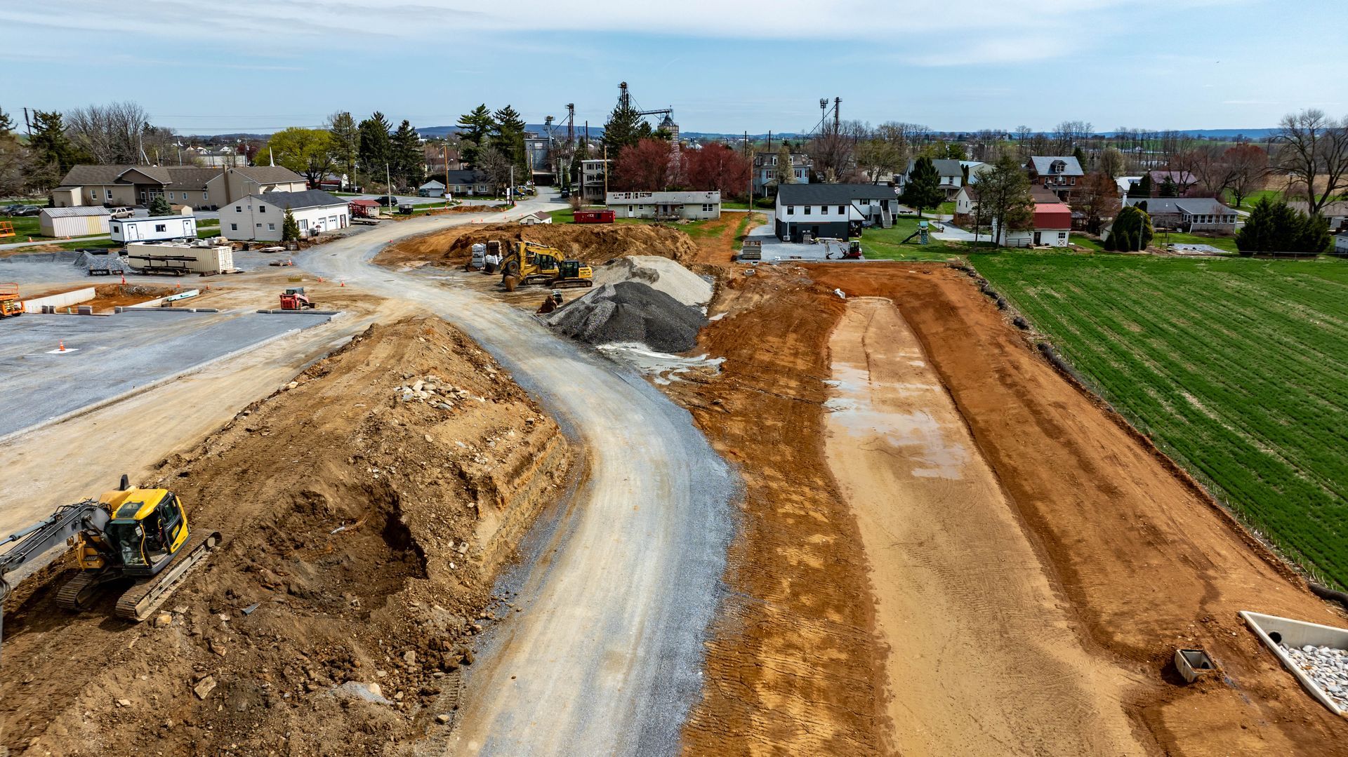 Construction site with excavators shaping a new road beside green fields and houses under clear skies