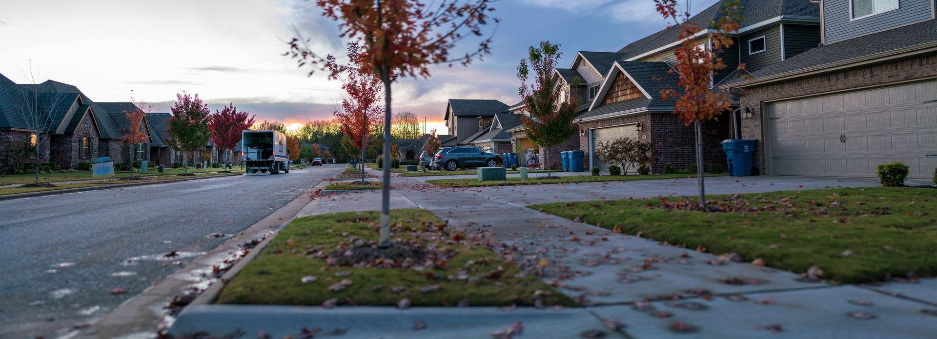 Quiet suburban street with houses, a sidewalk, and trees at dusk.