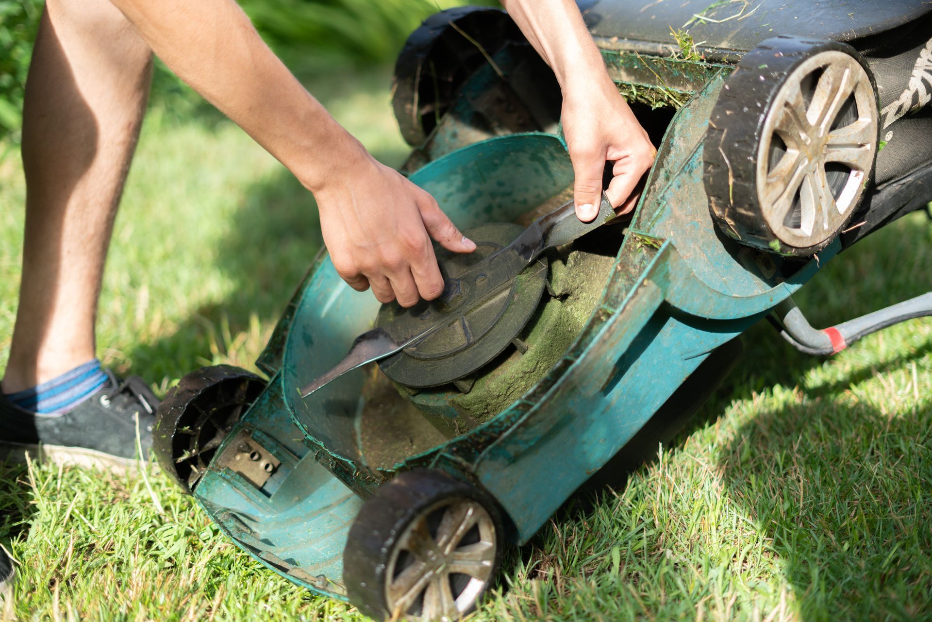 Person flipping a green lawn mower on grass, hands adjusting the underside by a wheel