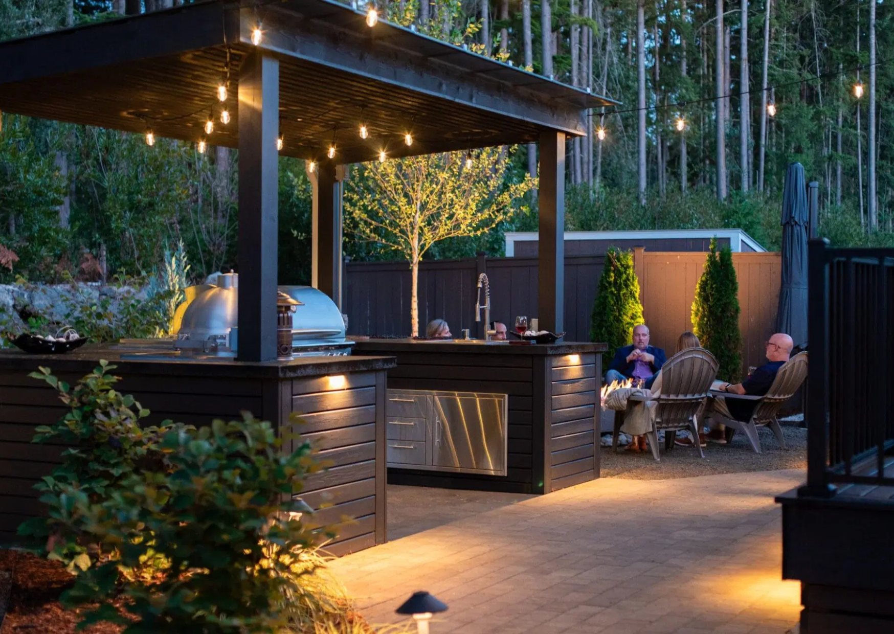 Modern outdoor kitchen in twilight with party guests in the background enjoying a firepit.