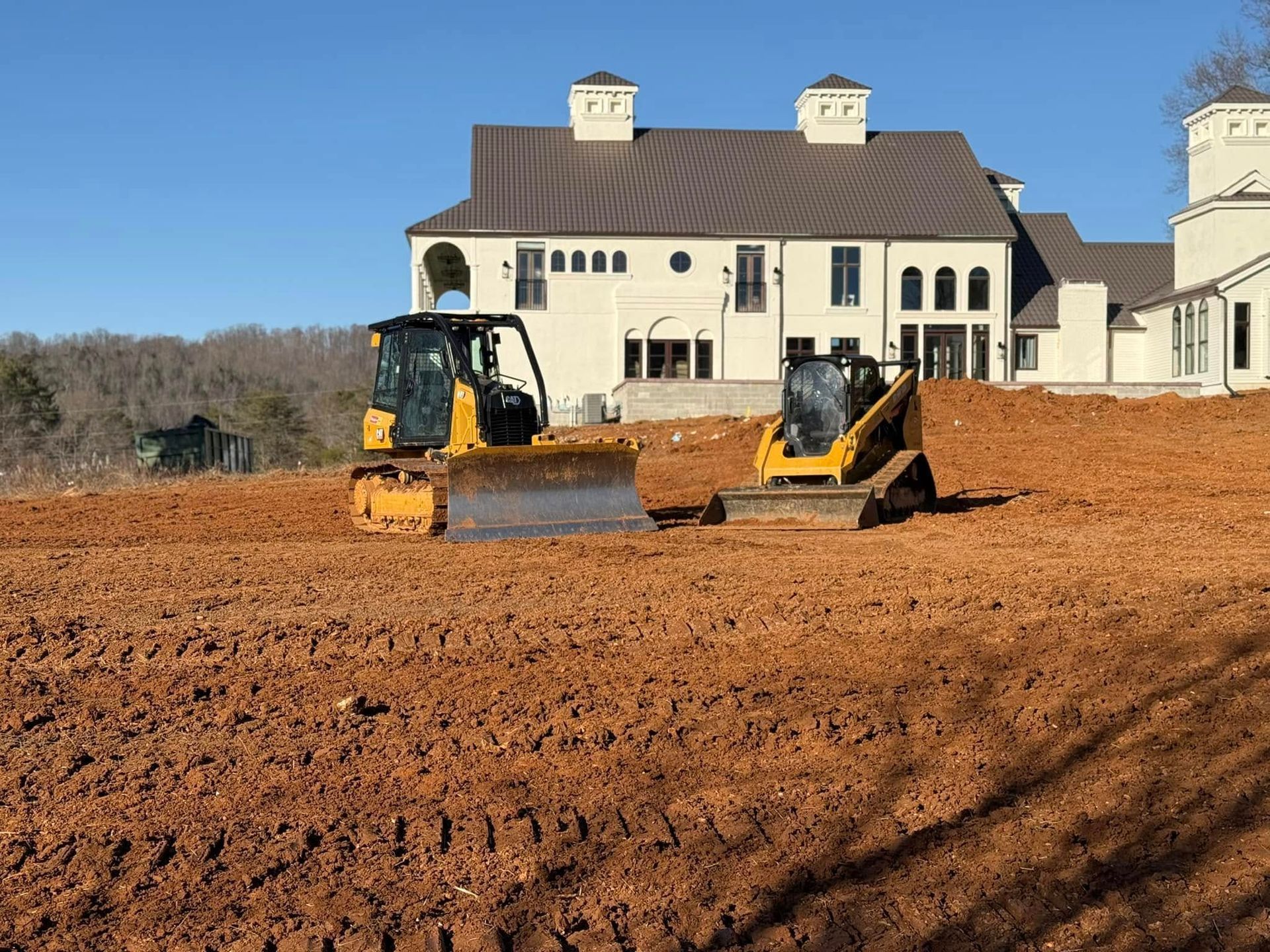 Two bulldozers are sitting in a dirt field in front of a large house.