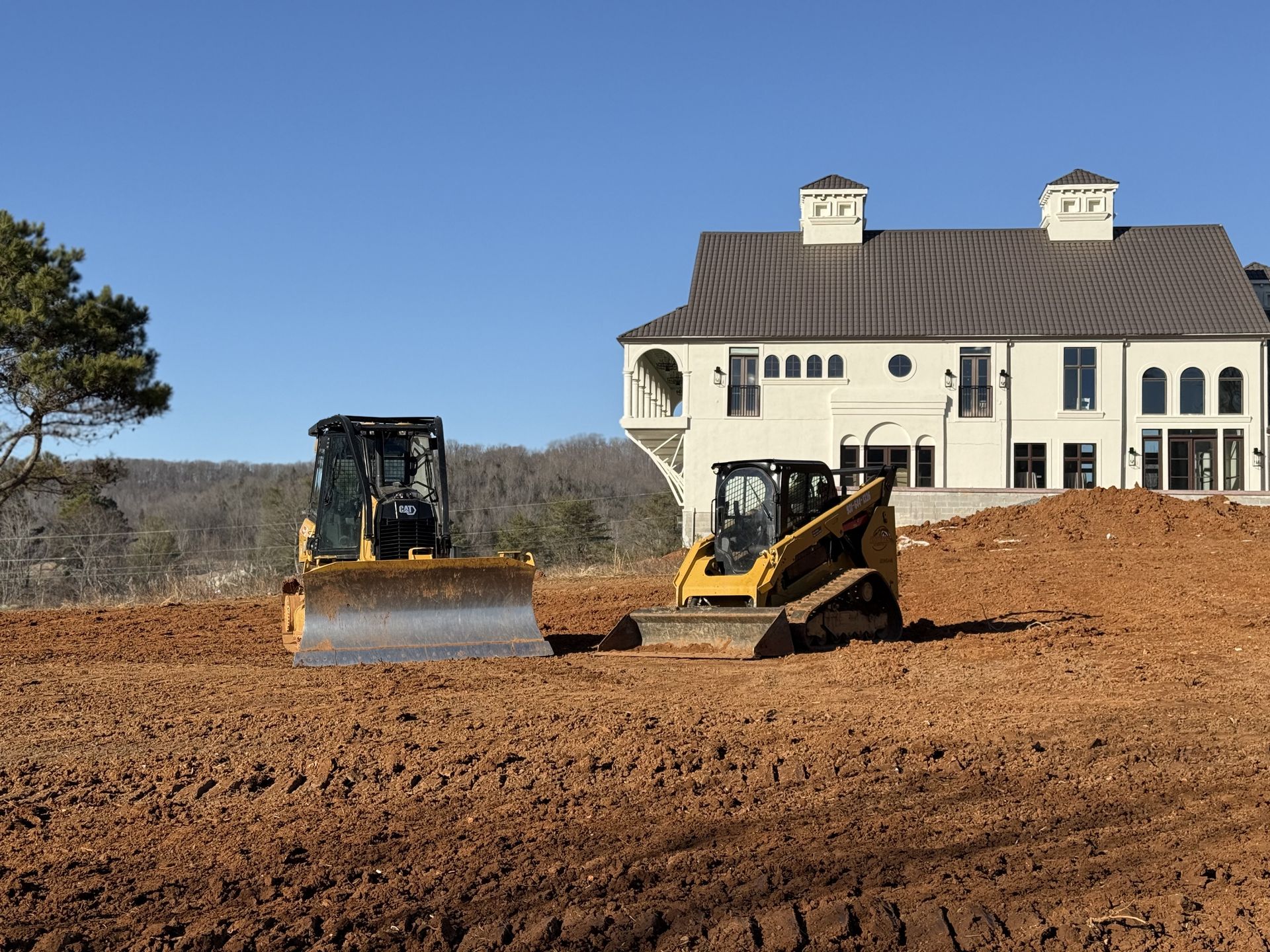 Two bulldozers are working in a dirt field in front of a large house.