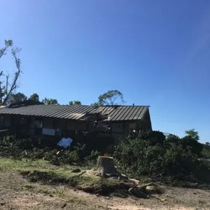 Damaged building with a partially collapsed roof, under a clear blue sky. Debris and cut tree stump visible.