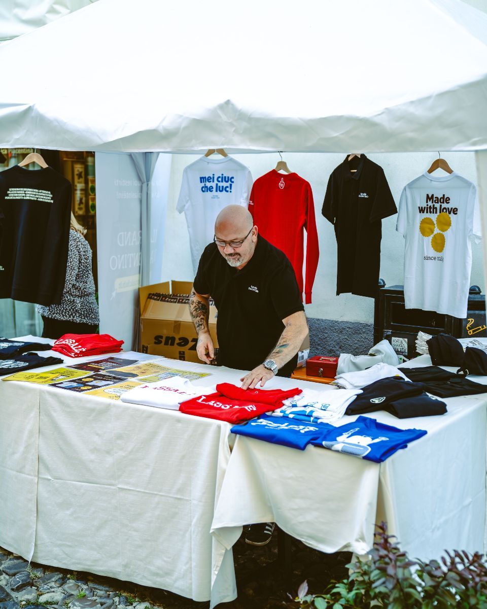 Uomo presso una bancarella che vende magliette. Le magliette sono esposte su un tavolo e appese sopra.