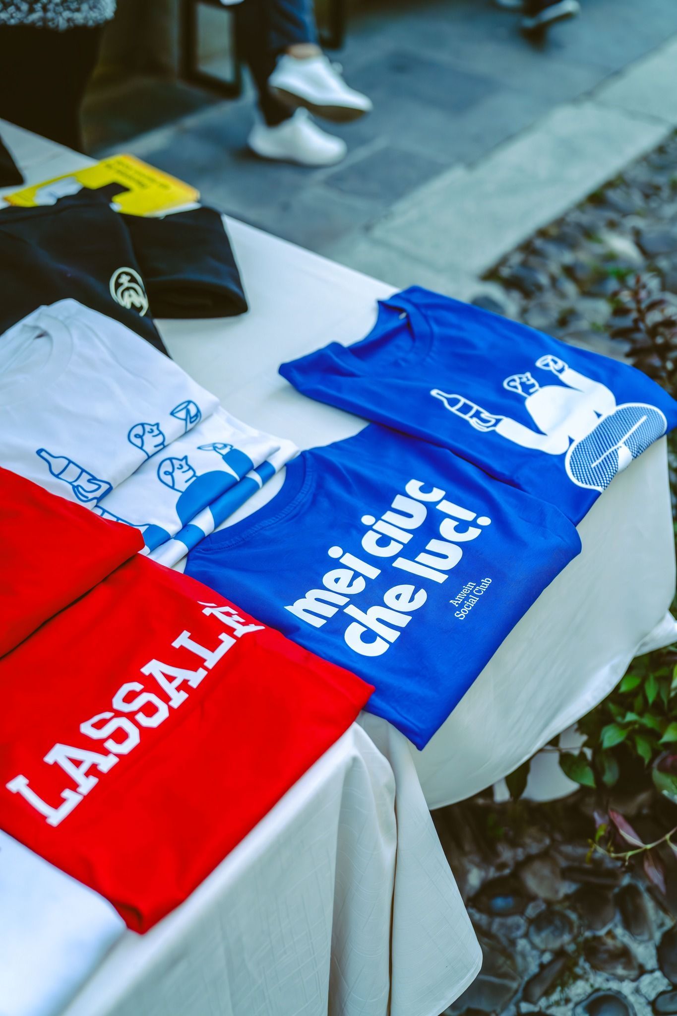 Magliette con scritte e disegni esposti su un tavolo. Sono visibili magliette rosse, bianche e blu.