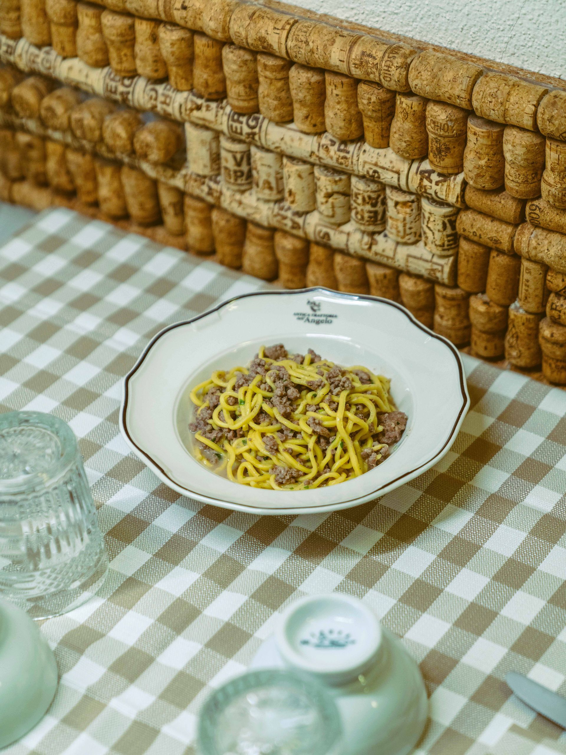 Piatto di pasta al ragù su tovaglia a quadri. Parete di sughero sullo sfondo.